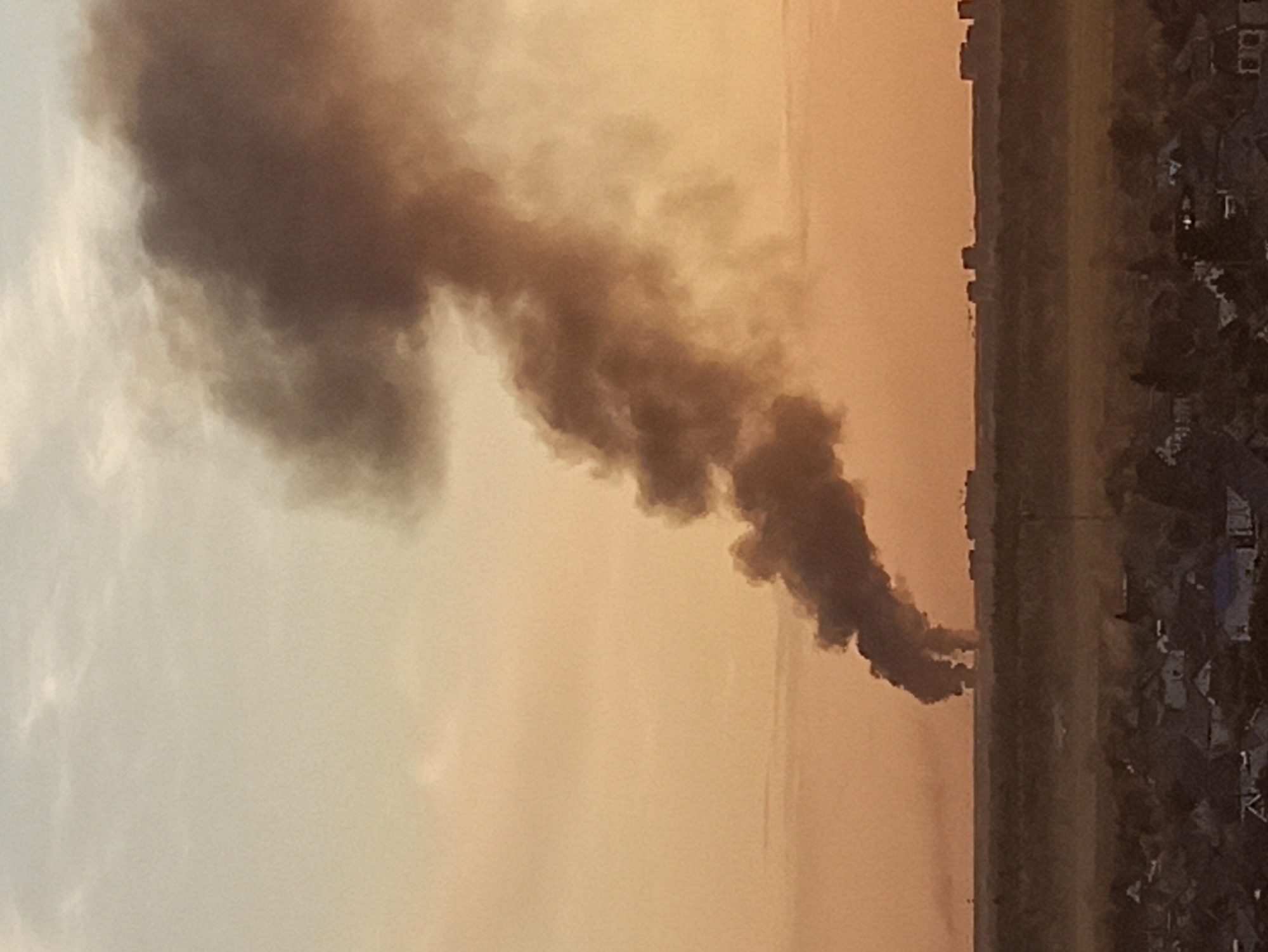 A plume of dark smoke rises into the orange sky near the village of Kondratovo, Russia. The source of the smoke appears to be on the edge of the village. The village contains single-family residences and taller buildings. The sky is overcast.