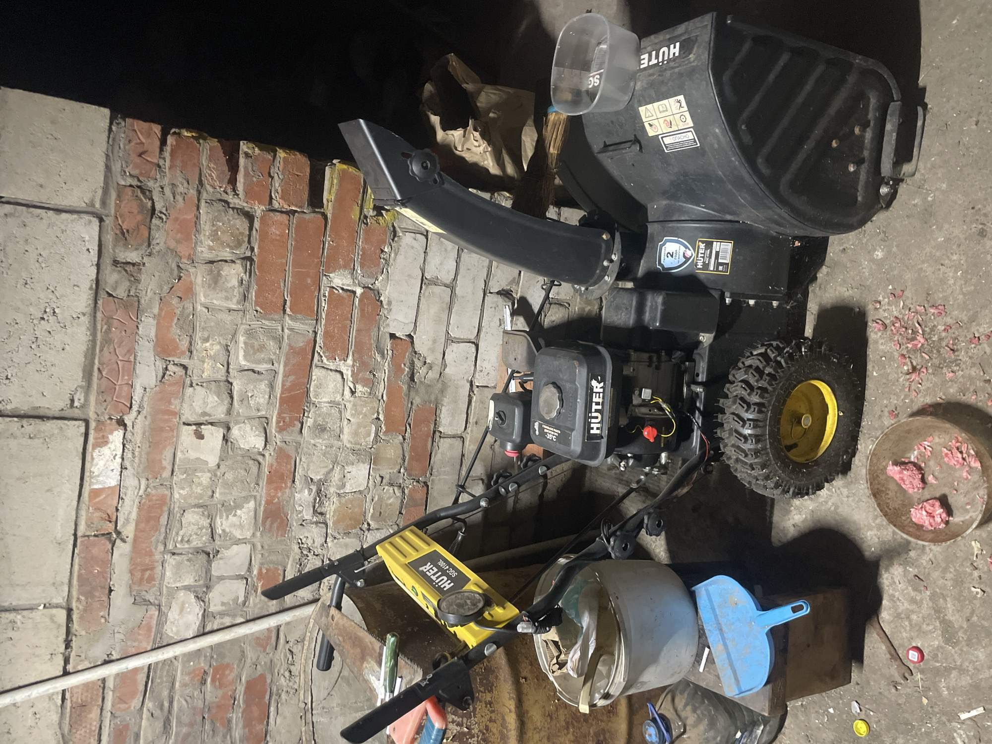 The image shows a dark, cluttered garage or storage area with a brick wall on the left. The primary object is a black Huter snowblower, positioned at an angle. It has a large chute and a yellow wheel with a black tire. Various tools and objects are scattered around the snowblower and on the floor, including a blue dustpan, a saw blade, and a small bowl containing some pinkish material. A brown paper bag is visible behind the snowblower. The lighting is dim, with shadows cast by the objects, suggesting it might be evening or nighttime. Some labels are visible on the snowblower, including "HUTER" and a blue sticker with a "2" and "ГОДА" (years in Russian). Other text appears to be warning symbols and possibly Russian language instructions. The scene is static, with no activity taking place. The location is identified as Laishevo, Russia.