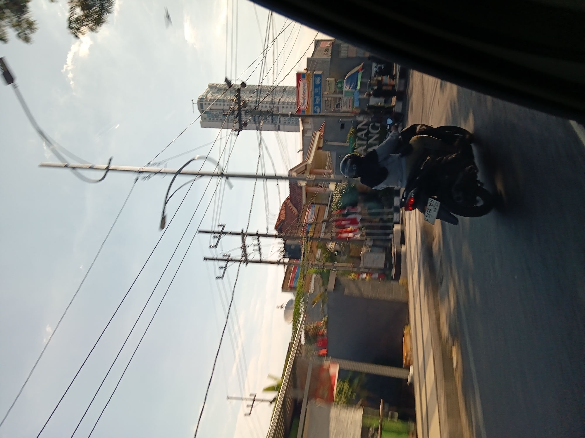 A motorcyclist wearing a black helmet and grey jacket is riding a black scooter down a street in Surabaya, Indonesia. The scooter has a license plate that reads "W 5286 YFU". In the background, there is a tall, modern building, a tall utility pole with wires, and several other buildings and shops. The sky is clear with some clouds. The image appears to be taken from inside a moving vehicle.