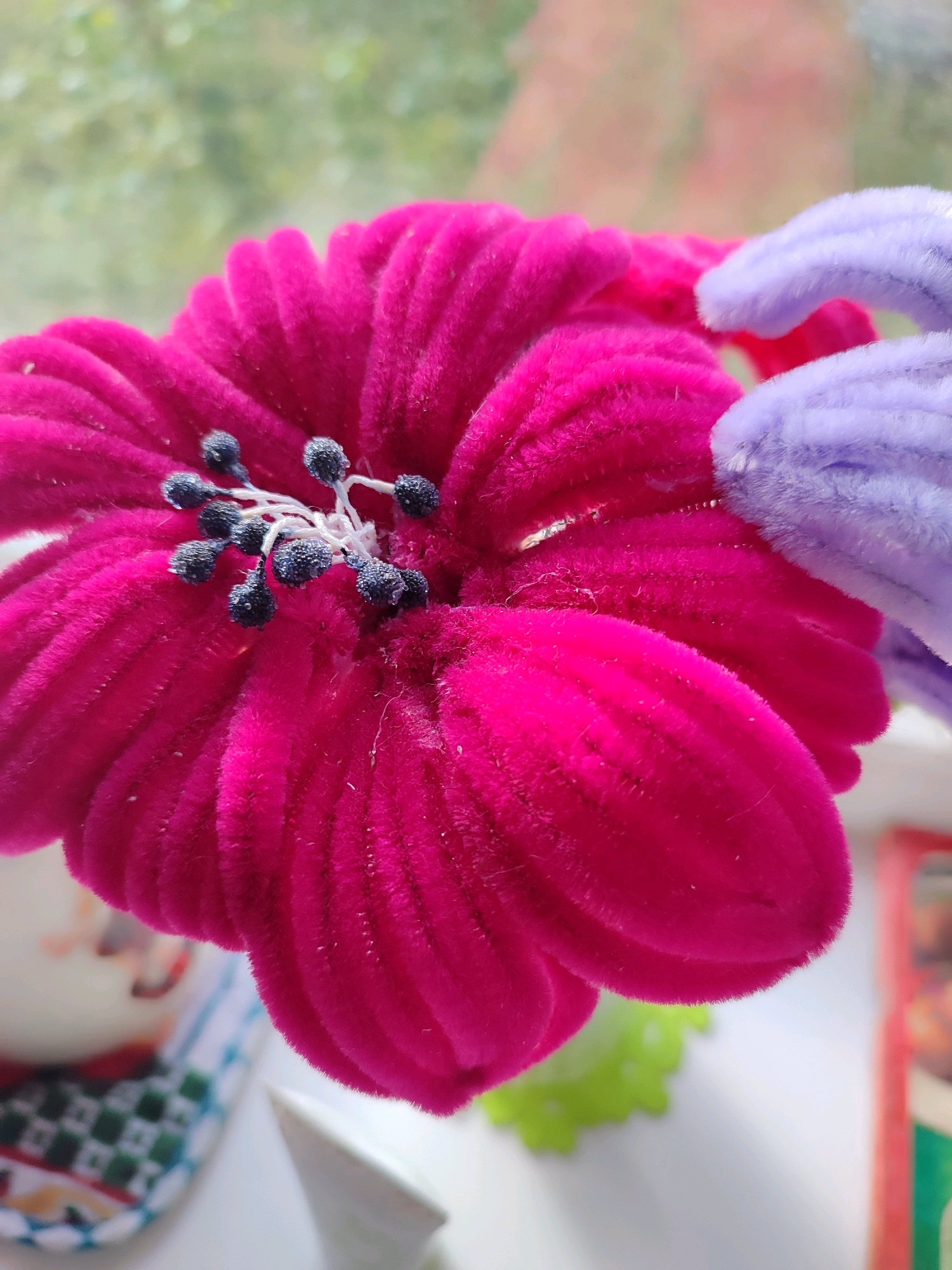A close-up, eye-level shot focuses on the center of a vibrant magenta, velvet-textured artificial flower. The flower has numerous petals radiating outwards, and its center features a cluster of stamens with dark blue, almost black, bead-like tips. The background is softly blurred, showing green foliage outside a window and the edge of a light-colored surface, possibly a windowsill. To the right and slightly behind the magenta flower, a fuzzy, lavender pipe cleaner curves into view, partially obscuring a portion of the magenta flower. The overall lighting is soft, suggesting an indoor setting near a window.