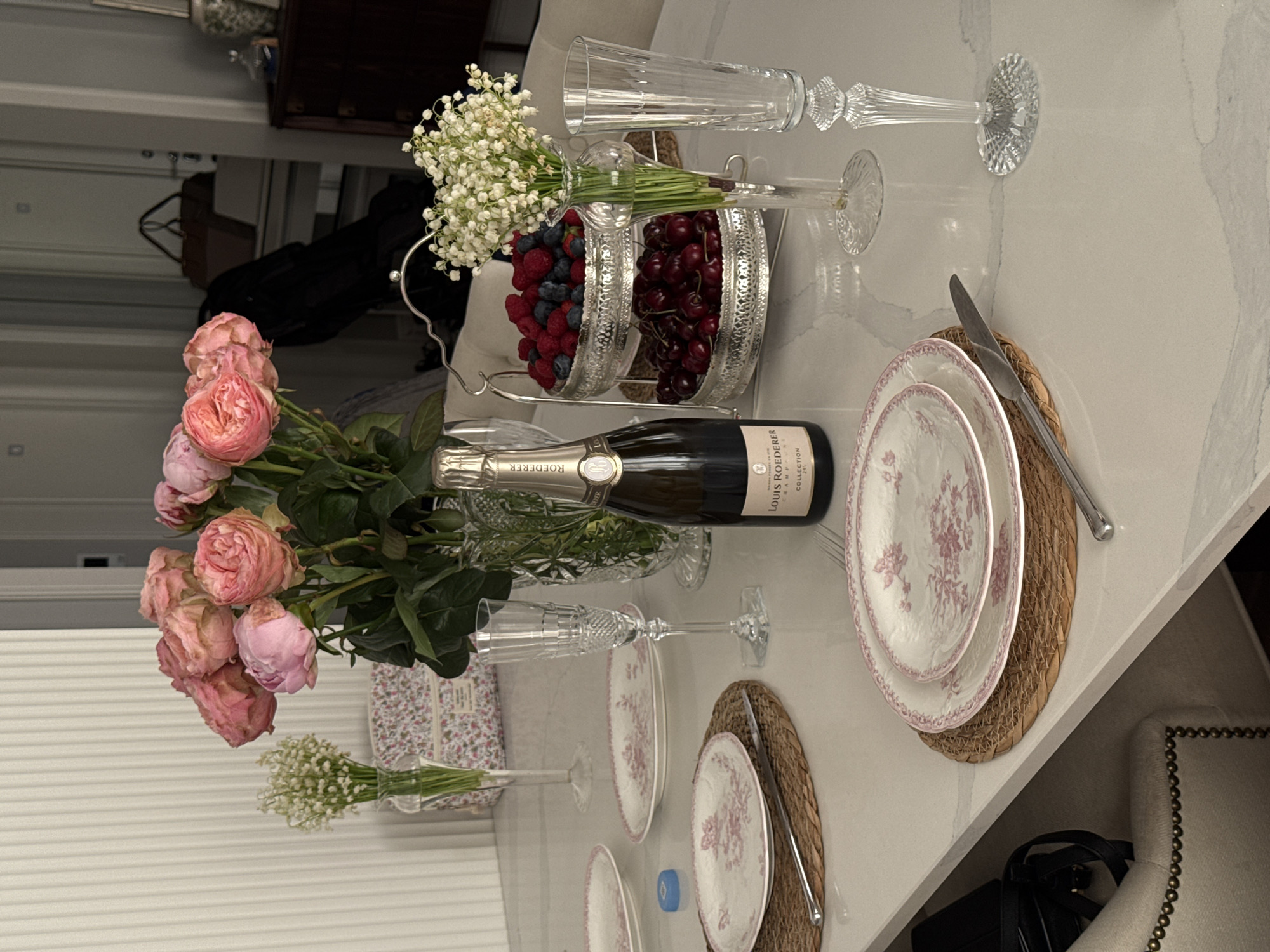 The image depicts a table setting with floral arrangements, tableware, and a bottle of champagne. There are pink roses and white flowers in vases, plates with a floral pattern on woven placemats, and champagne flutes. A silver serving dish contains cherries and raspberries. A bottle of Louis Roederer champagne is positioned near the flowers and plates. The scene appears to be indoors, likely in a dining room or kitchen. The location in Moscow, Russia, cannot be determined from the image alone.