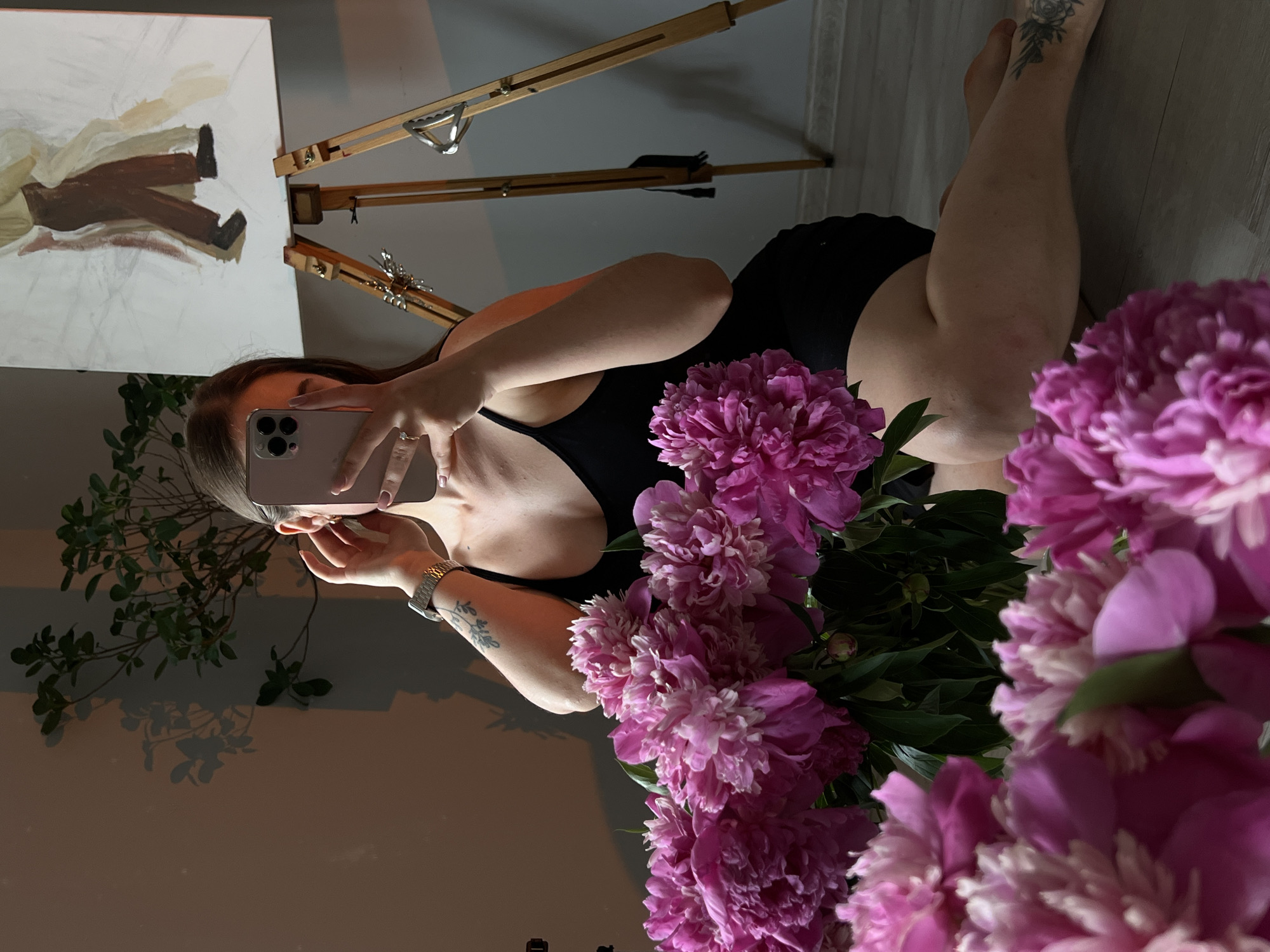 A woman is taking a selfie in a room with natural light casting shadows. She is wearing a black tank top and sitting on the floor with a bouquet of pink peonies in front of her. To her right, an easel holds a partially finished painting of two figures. The room appears to be a home or studio, with a plant visible in the background.
