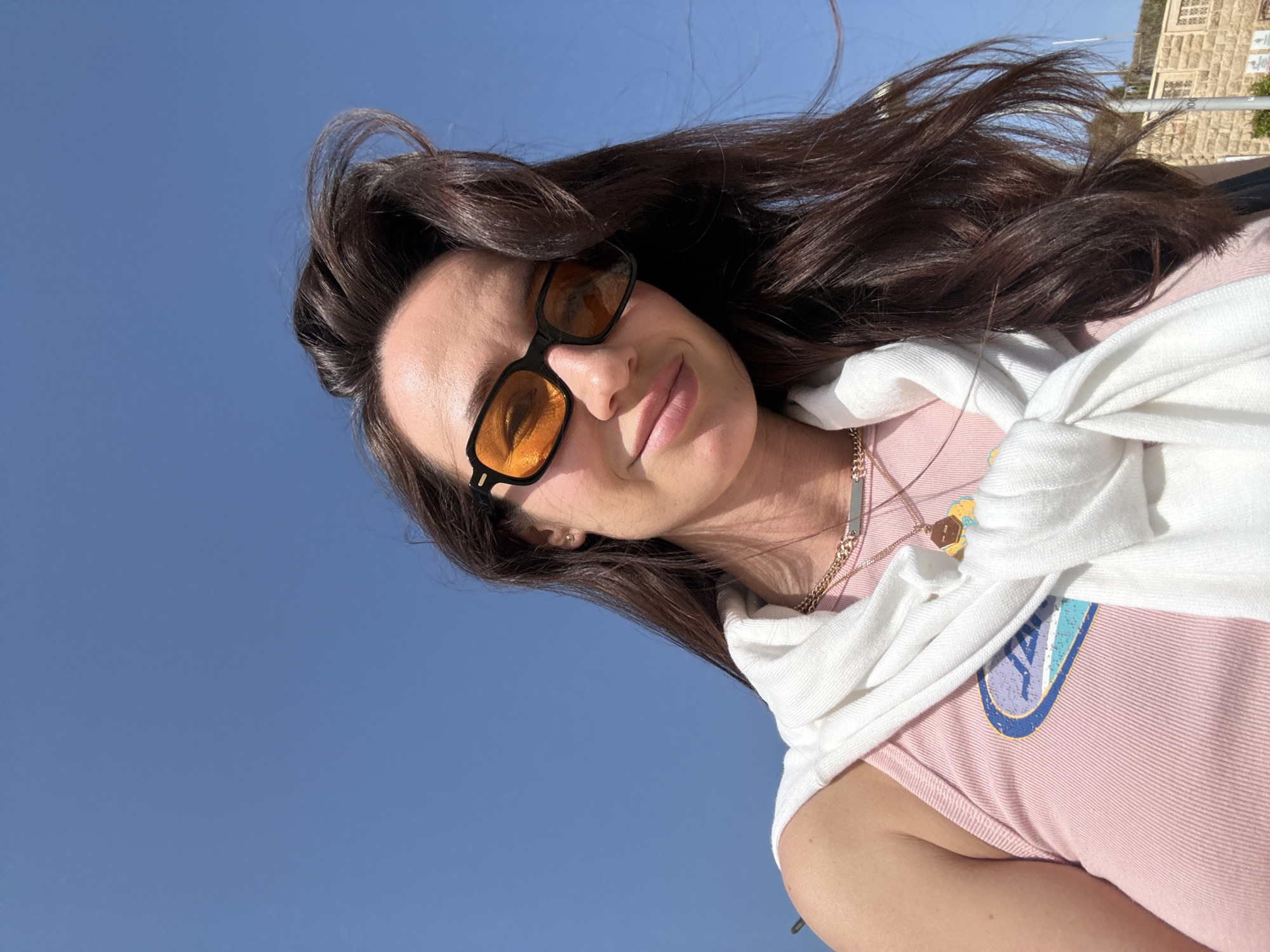 A woman with brunette hair, fair skin, and wearing sunglasses with amber-tinted lenses takes a selfie outdoors in Tel-Aviv, Israel. She is wearing a pink t-shirt and a white sweater draped around her shoulders. A multi-layered necklace is visible. The background includes a clear blue sky and a stone building in the distance. The woman's hair is windblown.