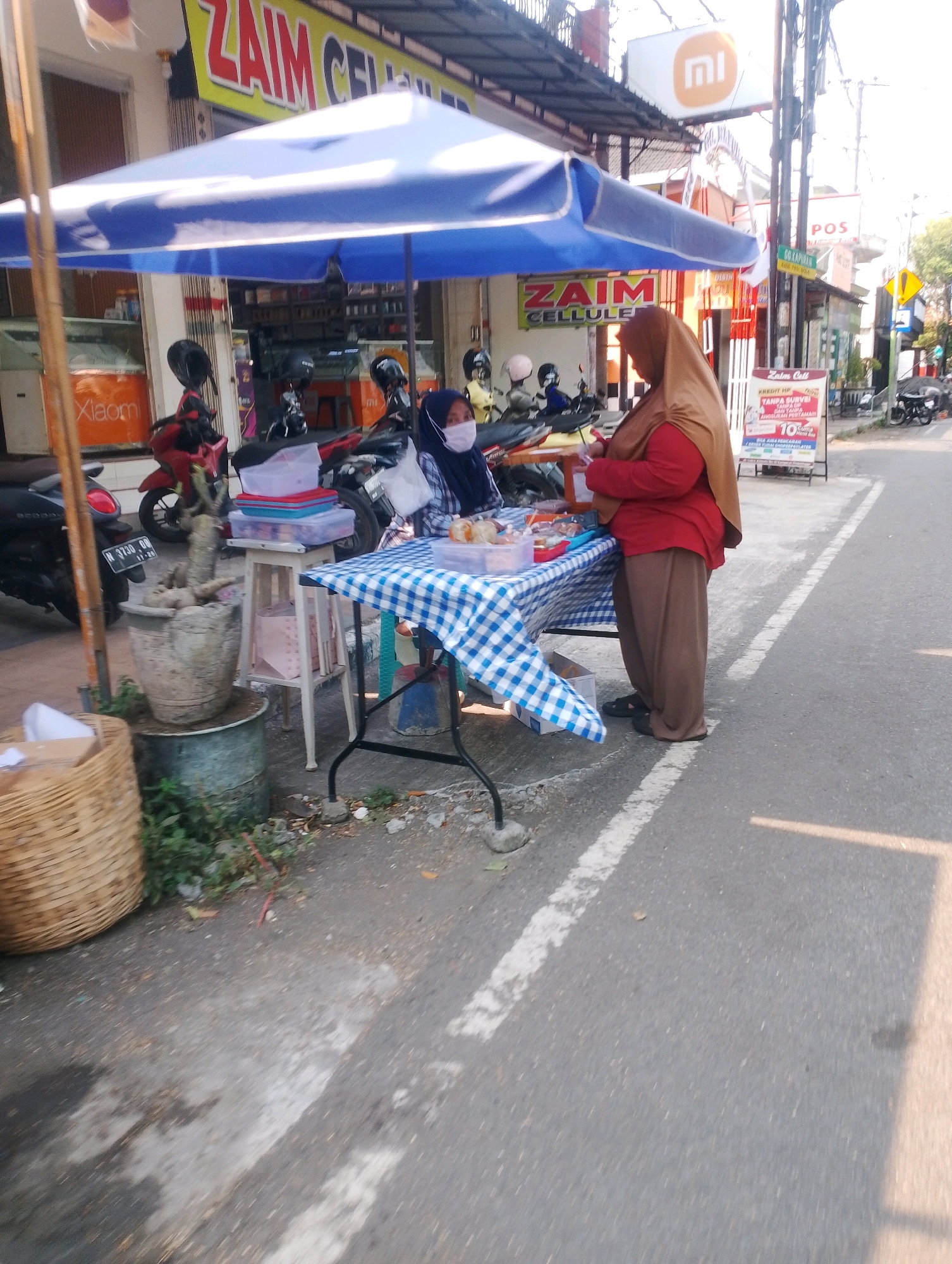 In Probolinggo, Indonesia, a street-side vendor is seen selling goods from a table covered with a blue and white checkered cloth. The vendor, wearing a brown hijab and a red shirt, stands behind the table, attending to customers or organizing her merchandise. Another person, partially obscured and wearing a blue hijab and a mask, sits at the table, possibly a customer or helper. The table displays various items, including food items in clear containers and stacked plastic boxes. Behind the vendor, a sign reads "ZAIM CELLULE," suggesting the establishment is a mobile phone or telecommunications shop. The scene takes place outdoors on a paved road, with motorcycles parked nearby, indicating a busy urban or commercial area. The environment appears to be a sunny day.