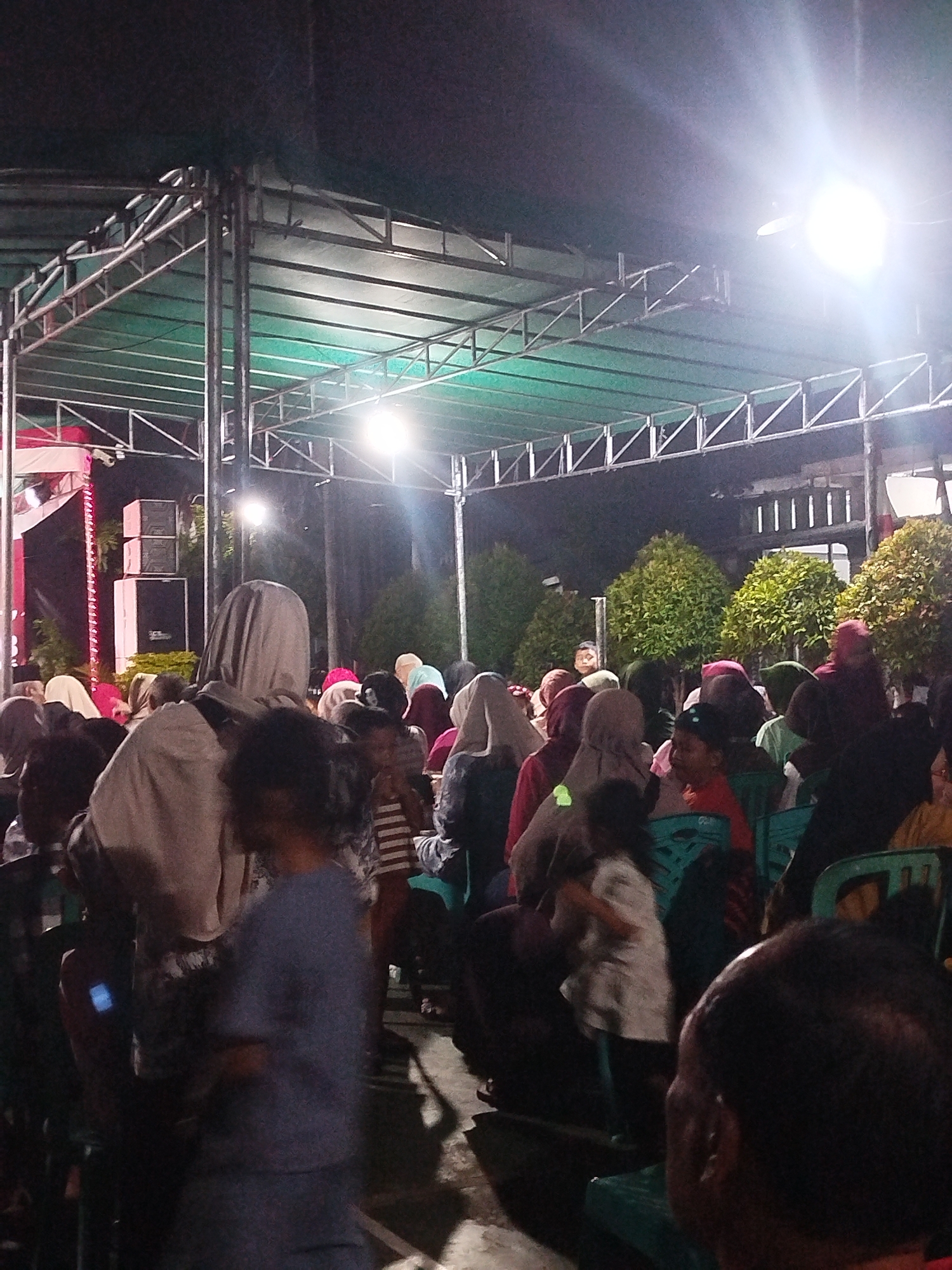A crowd of people, many wearing hijabs, are gathered outdoors at night under a green canopy supported by metal scaffolding. The scene appears to be a community event or gathering. Stage lighting illuminates the area from the right, creating bright spots and casting shadows. In the background, there are trees and buildings. On the left side of the frame, a stage with red and white draping and stacked speakers is visible. The overall atmosphere suggests a lively, albeit somewhat dimly lit, public event in Depok, Indonesia.