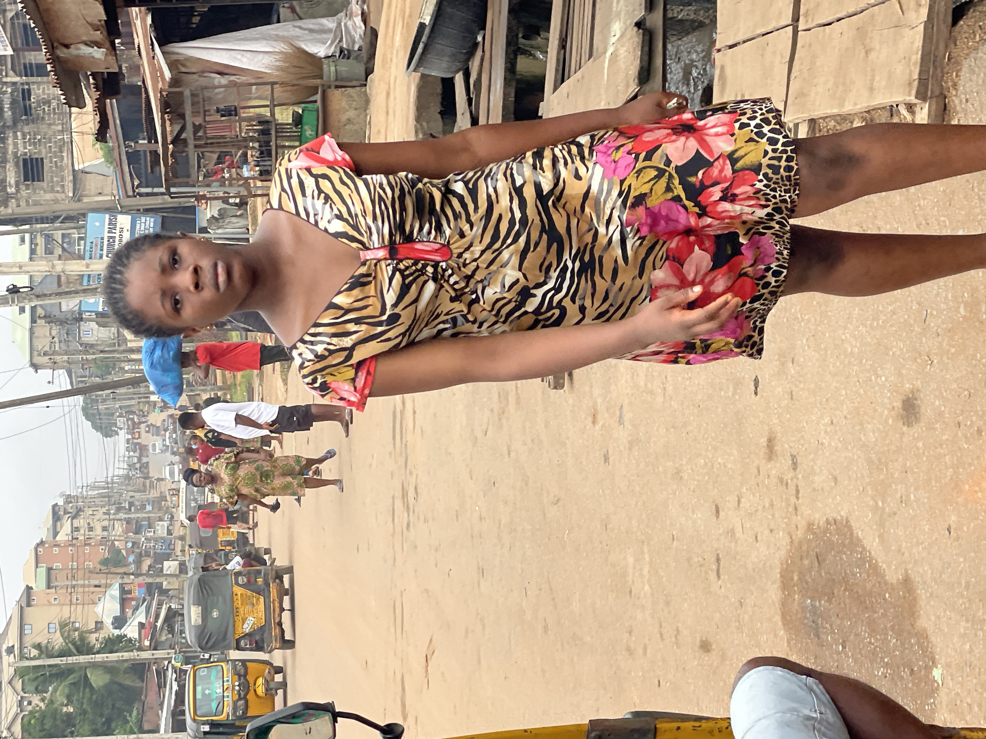 The image depicts a street scene in Nkpor, Nigeria. In the foreground stands a young woman wearing a short, patterned dress with a tiger stripe and floral design. Behind her, a street stretches into the distance, populated with people, vehicles (primarily auto rickshaws, also known as "keke"), and buildings. The buildings are a mix of residential and commercial structures, with varying heights and architectural styles, typical of an urban environment in Nigeria. The street surface is paved but appears worn and possibly uneven. The sky is overcast, contributing to a somewhat muted lighting in the scene. The woman is the main focus, standing out due to her attire and central positioning.