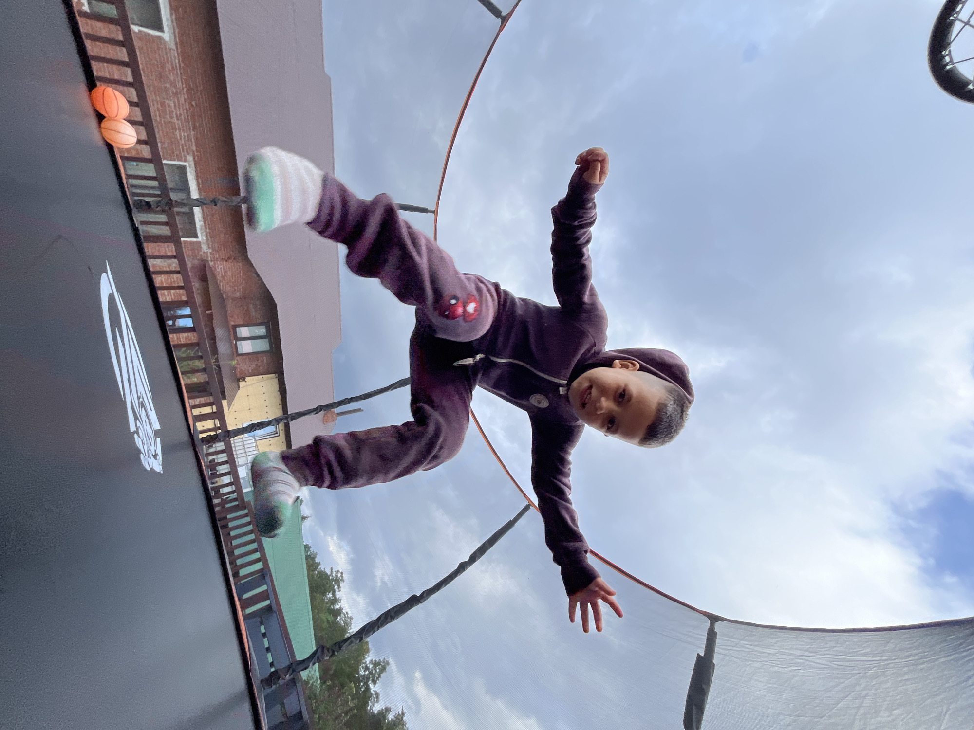 A young boy is captured mid-air, jumping on an outdoor trampoline. He is wearing a dark purple tracksuit with colorful patches on the knees and striped socks. The trampoline is black with a white logo and is surrounded by a safety net. In the background, there is a residential building with a red brick facade and a green roof, along with trees. Two small orange balls are visible on the edge of the trampoline. The sky is cloudy and overcast. The image appears to be taken from a low angle, looking up at the boy. The setting is likely a backyard in Chaikovsky, Russia, given the architectural style and foliage.
