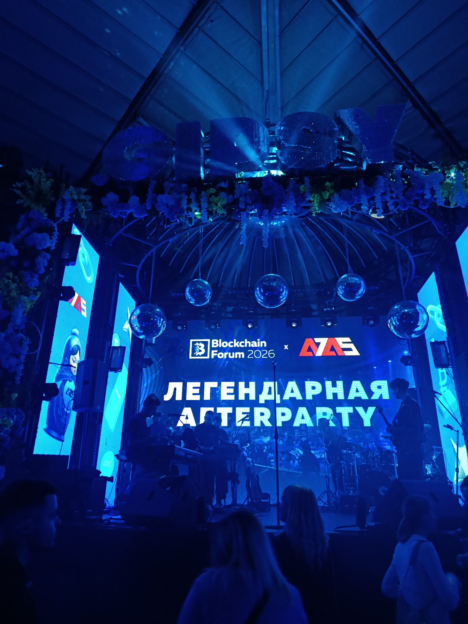 The image shows a stage at an event, possibly a conference or afterparty, located in the city of Unknown, Unknown. Above the stage, large, illuminated letters spelling "JOY" are visible. The stage itself is decorated with flowers and foliage. The backdrop features a large screen displaying the logos for "Blockchain Forum 2026" and "AZAS," along with the Russian text "ЛЕГЕНДАРНАЯ AFTERPARTY" (Legendary Afterparty). A band is performing on stage, and several disco balls hang above them. Attendees are visible in the foreground, watching the performance. The scene is bathed in a blue light.