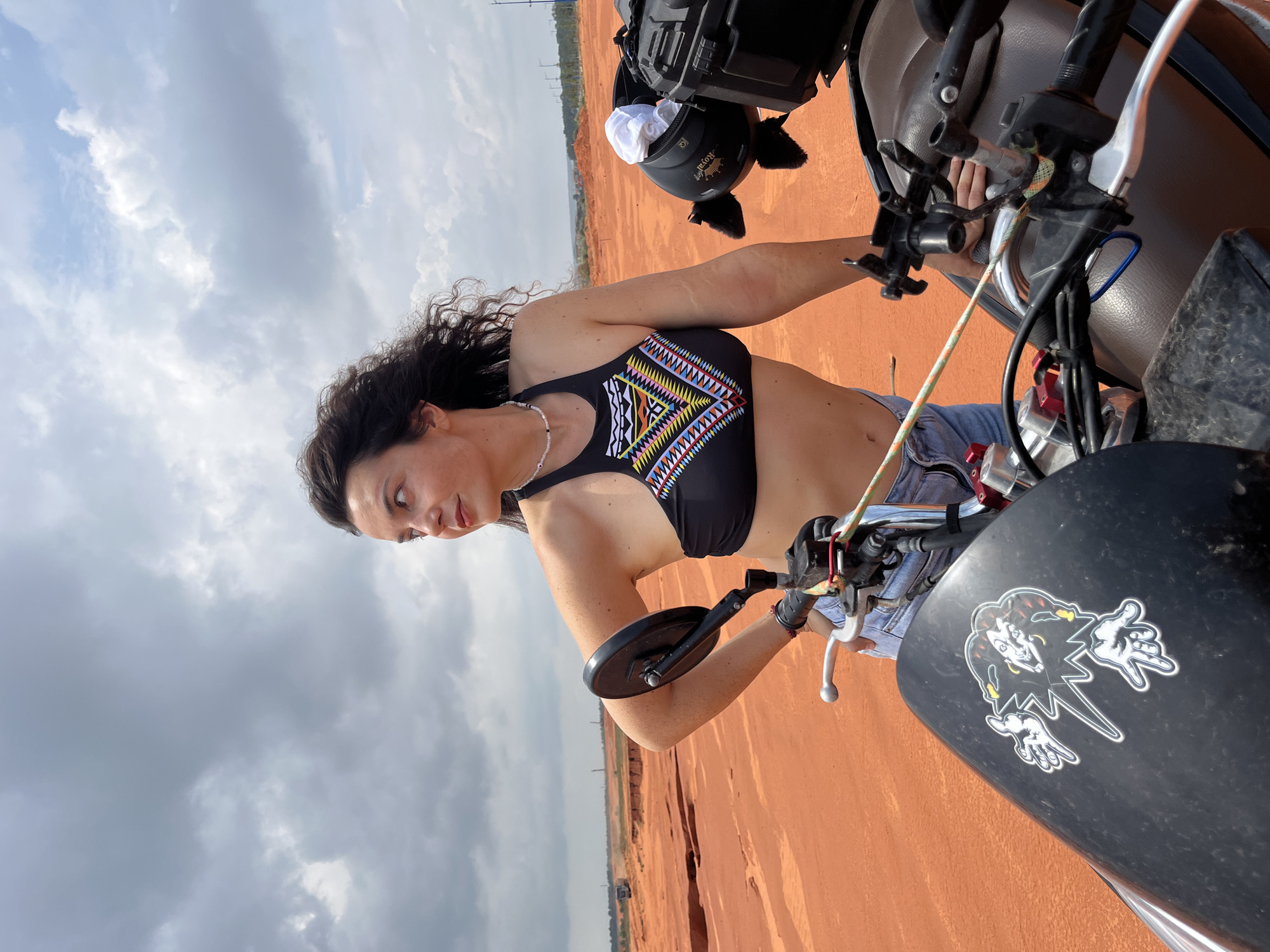 In Mui Ne, Vietnam, a woman stands next to a motorcycle in a sandy, construction-like area under a cloudy sky. She has long, dark, curly hair and is wearing a black bikini top with a colorful, geometric pattern and denim shorts. Her left hand is on her hip, and her right arm is resting on the motorcycle's seat. The motorcycle has a black windshield with a sticker of a clown-like figure, and a black storage box is mounted behind the seat, containing a white helmet. The ground is a reddish-orange color, typical of the sand dunes in the region. In the background, distant trucks are visible, suggesting ongoing construction or development. The overall impression is one of a rugged, outdoor setting.