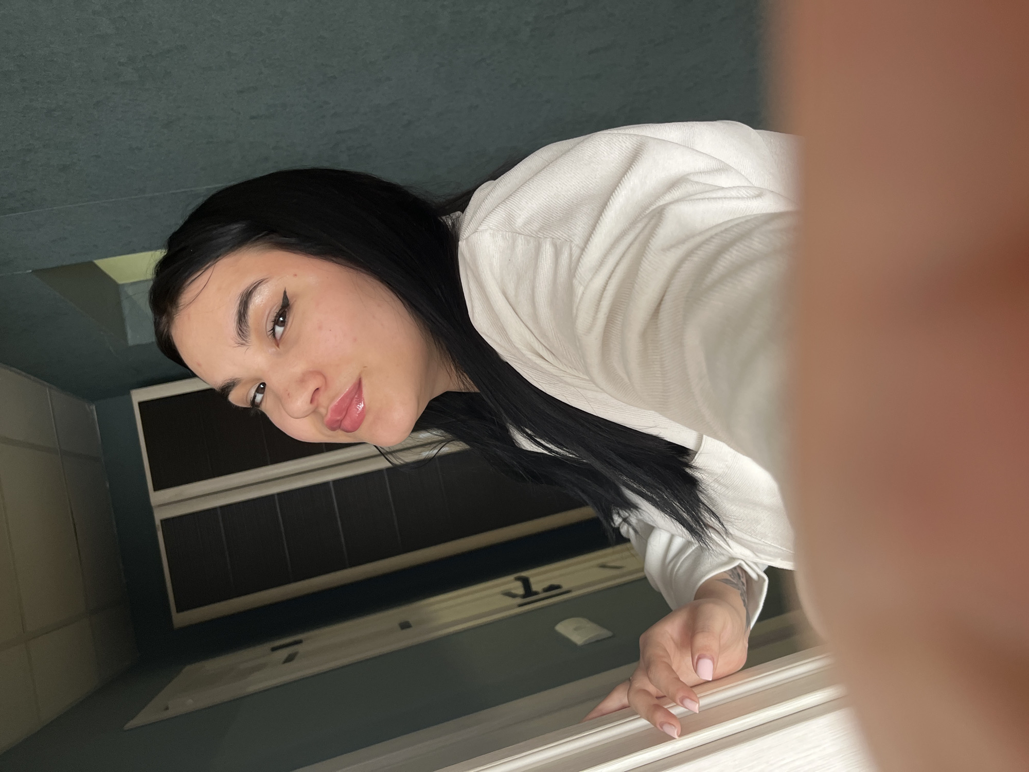 A young woman with dark hair and prominent winged eyeliner is depicted in a selfie, smiling directly towards the camera. She wears a light-colored, possibly white or cream-colored, long-sleeved ribbed top. Her right hand, featuring light pink manicured nails and a faint tattoo on the forearm, rests on a white door frame on the left side of the image. The background reveals an interior hallway or room with dark teal-colored textured walls. A white panel ceiling is visible at the top. To the left, a partially open white door with a dark handle is seen, along with a set of darker framed double doors further in the background. The perspective is a close-up, low-angle shot, with the camera-holding hand partially obscuring the bottom right. The scene is described as being in Chaikovsky, Russia.