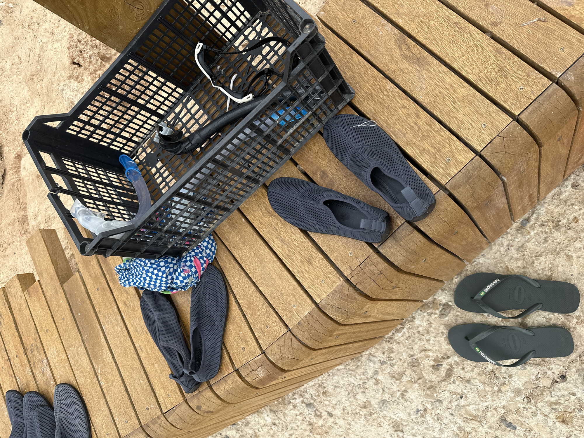 An outdoor scene shows a collection of footwear and snorkeling gear arranged on a wooden bench and the sandy ground. The bench is made of curved, light brown wooden planks with visible grain and some wear. On the bench, a black plastic crate contains a blue and white patterned fabric item and snorkeling equipment, including a snorkel and mask. Several pairs of dark grey water shoes are also present, some on the bench and some on the ground. A pair of dark grey flip-flops are on the sandy ground in the foreground. The environment appears to be a beach or poolside setting with sand and a concrete-like surface visible. The overall scene depicts items prepared for or just used for water activities.