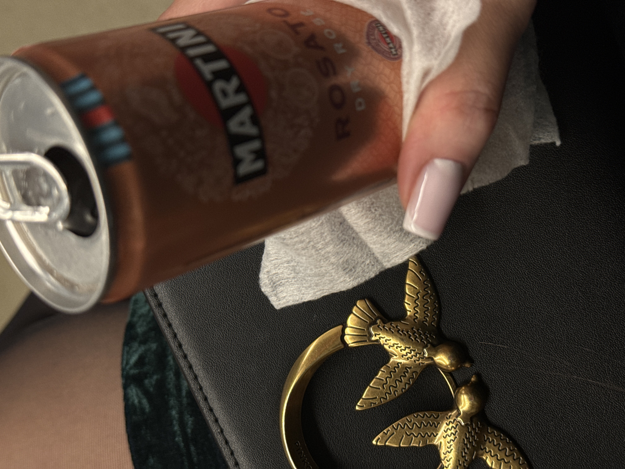 The image shows a partially visible can of Martini Rosato dry rosé sparkling wine held by a hand with a white nail polish design. A white tissue surrounds the can. Beneath the can, a black bag with a gold-colored bird-shaped clasp is visible. Part of a green velvet-like surface is also in the frame. The location in the city of Moscow, Russia cannot be determined from the visual information available.