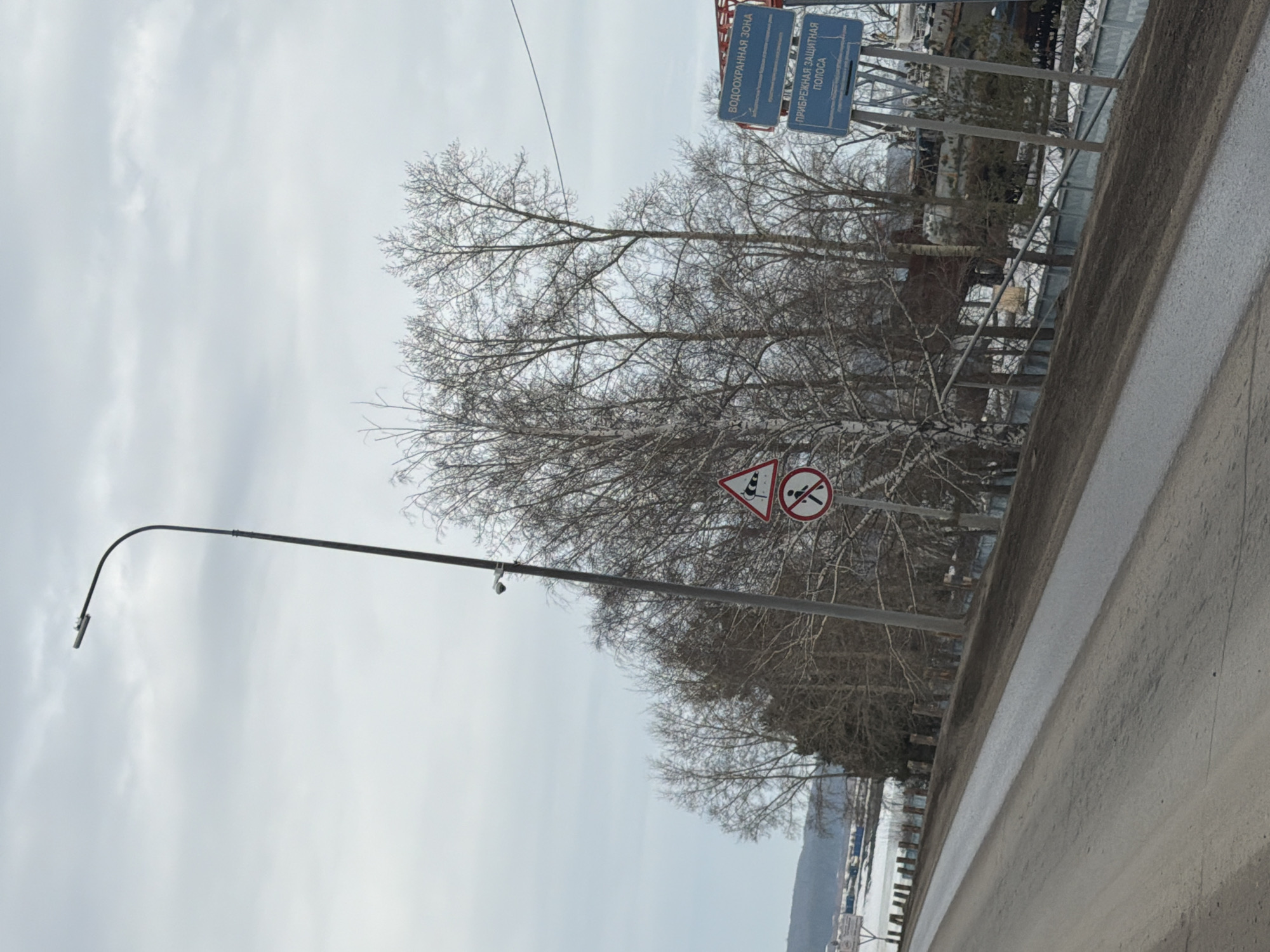 The image depicts a view of the city of Chaikovsky, Russia, alongside a road with a concrete barrier. A street light with a surveillance camera extends from the left side. In the background, there are bare trees and signs, including those indicating a water protection zone. A sign with a white triangle with a dark border and a dark exclamation mark in the middle of it along with a sign with a red circle, a diagonal red line and a dark figure of a swimmer. The weather appears to be overcast. A body of water can be glimpsed at the bottom of the image.