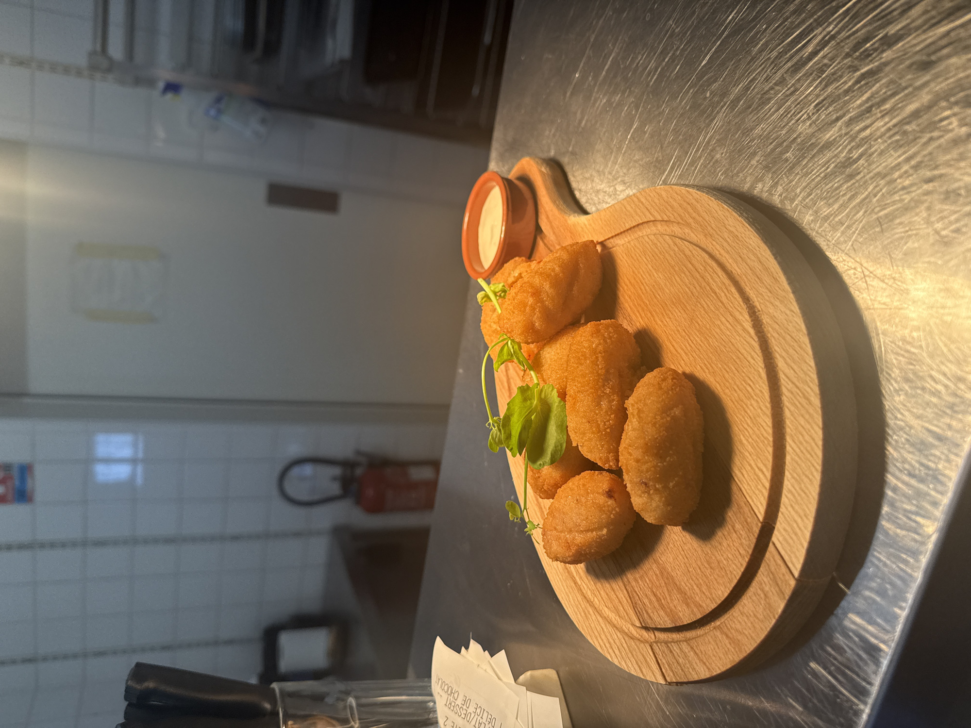 A close-up shot shows a wooden platter with several golden-brown croquettes, garnished with a sprig of green herbs. A small orange dish filled with a white dipping sauce sits next to the croquettes. The platter rests on a metallic surface, likely a countertop. In the background, a tiled wall, a fire extinguisher, and a metal shelving unit are visible. A crumpled receipt is partially visible on the left side of the image. The scene appears to be in a restaurant kitchen or service area in Rivesaltes, France. There are no people visible in the image.