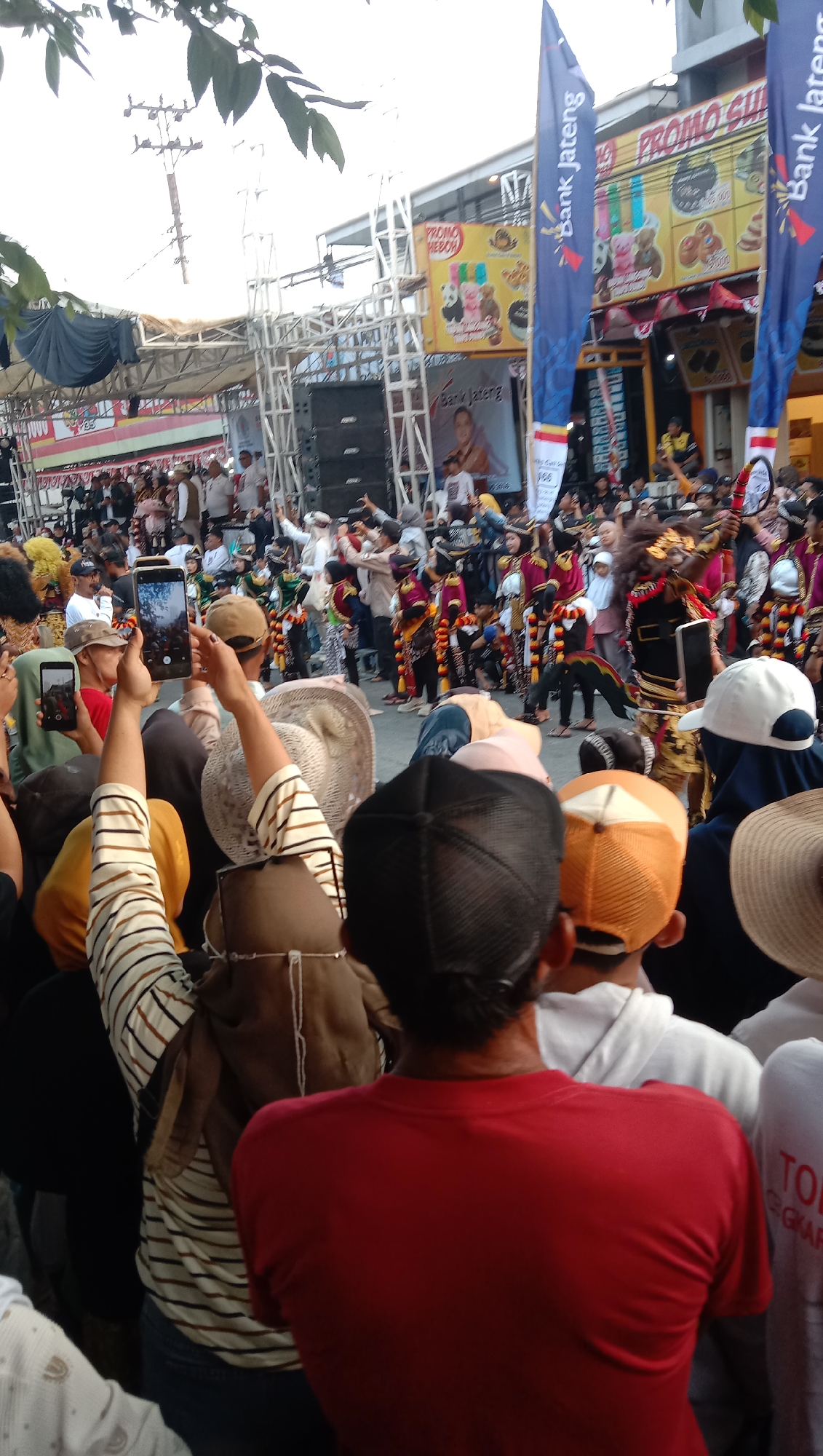 In Jatisawit, Indonesia, a lively street event is captured from behind a crowd of spectators. A group of performers in traditional colorful costumes, some wearing elaborate masks and headdresses, are visible on a stage or designated performance area. The crowd, densely packed, is actively engaged, with many individuals holding up their mobile phones to record the event. Some people in the foreground are wearing casual attire and hats, their backs to the camera, as they watch the performance. In the background, banners with Indonesian text, including "PROMO HEBOH" and "Bank Jateng," indicate commercial sponsorship. The scene suggests a cultural festival or public celebration taking place in a semi-urban or town setting.