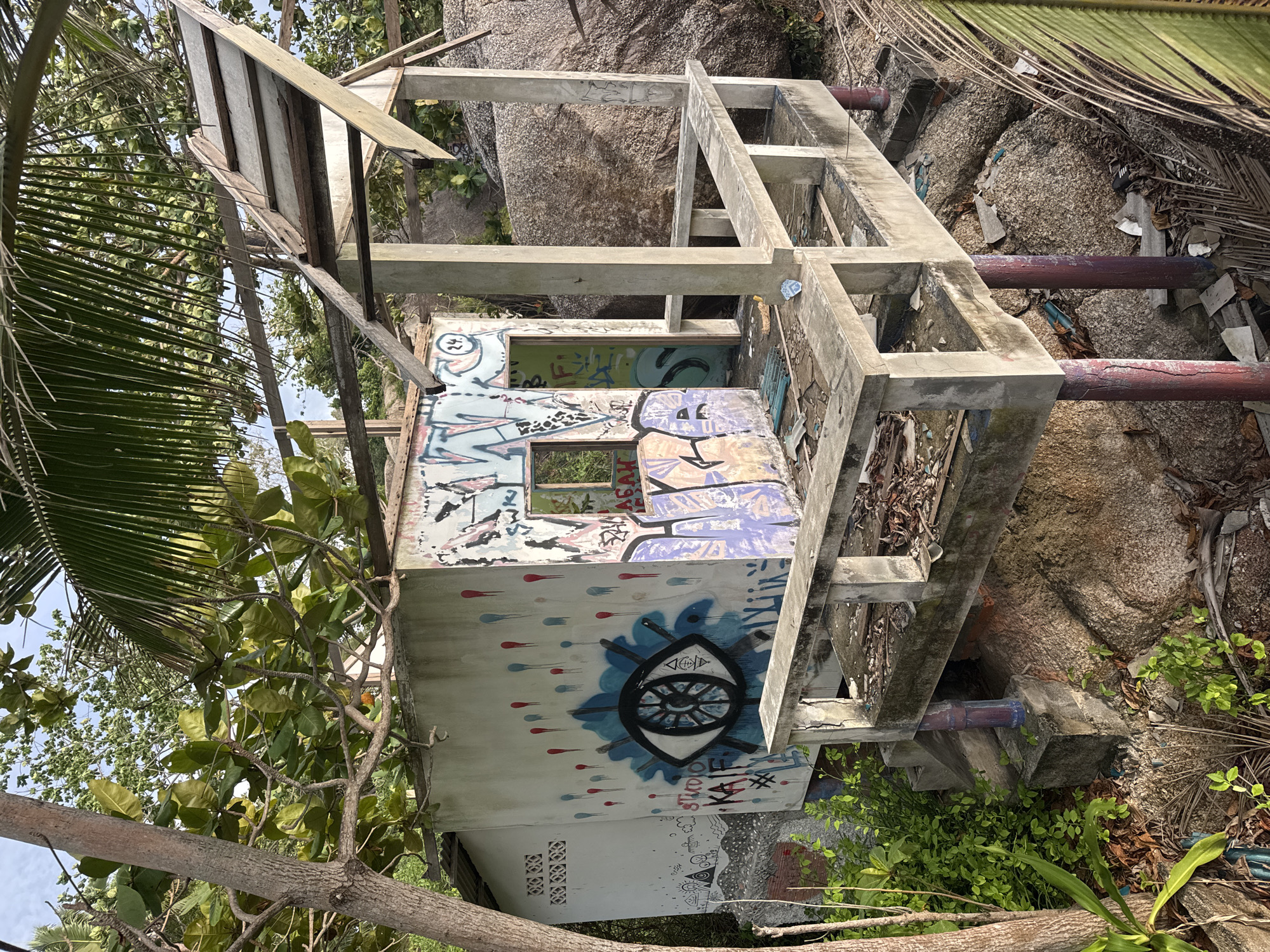 The image shows a dilapidated, possibly abandoned, concrete structure in Baan Thong Nang, Thailand. The structure has a small, graffiti-covered room with windows and is surrounded by a crude concrete framework that suggests it may have been intended to be multi-level. The graffiti on the room includes an eye design and other abstract patterns and lettering. The structure is located among rocks and foliage, indicating an outdoor setting, likely near the coast or in a rocky area. Several pipes also are seen nearby. The location appears to be in a state of disrepair and neglect. No people are visible in the scene.