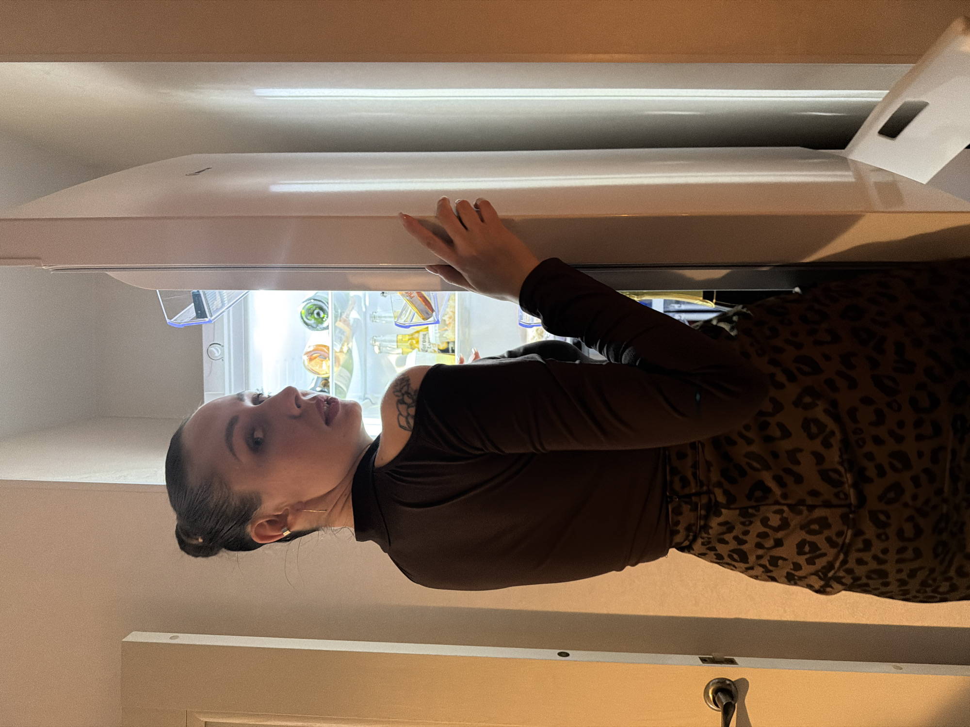 A woman is standing in front of an open refrigerator, looking into it. She has her left hand resting on the refrigerator door. She is wearing a dark brown long-sleeved shirt and a brown leopard print skirt. The refrigerator is well-lit, containing various items, including bottles, jars, and other food items. The environment appears to be a kitchen. There is no evidence to suggest the location is in Moscow, Russia.