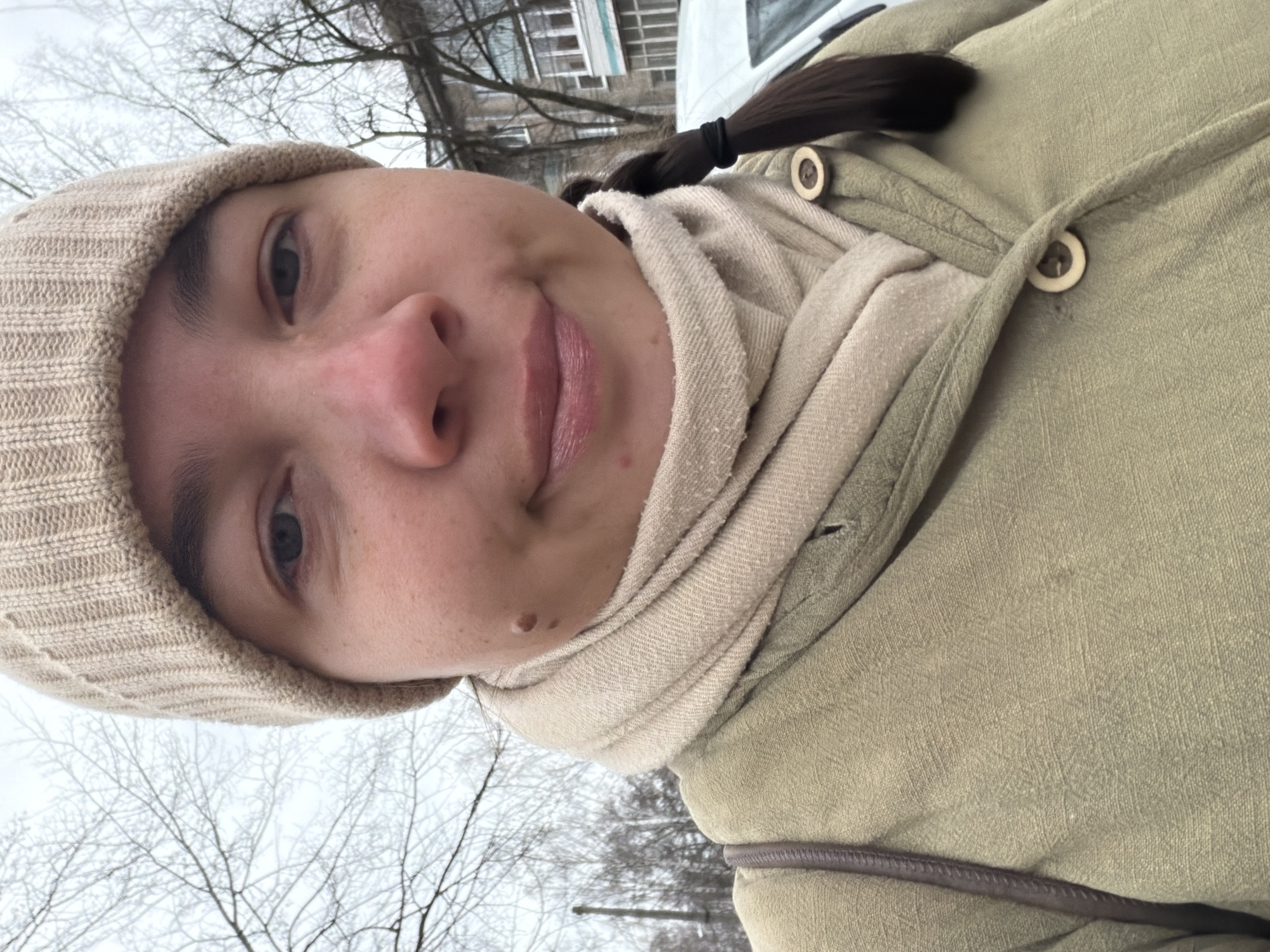 A close-up, slightly angled selfie of a person with fair skin, wearing a beige knit cap and a light-colored scarf. The person has dark hair pulled back into a ponytail. They are wearing an olive-green coat with wooden buttons. In the background, bare trees and a light-colored building are visible. The location is likely outdoors, given the trees and overcast sky. The image does not provide enough information to confirm it was taken in Chaikovsky, Russia.