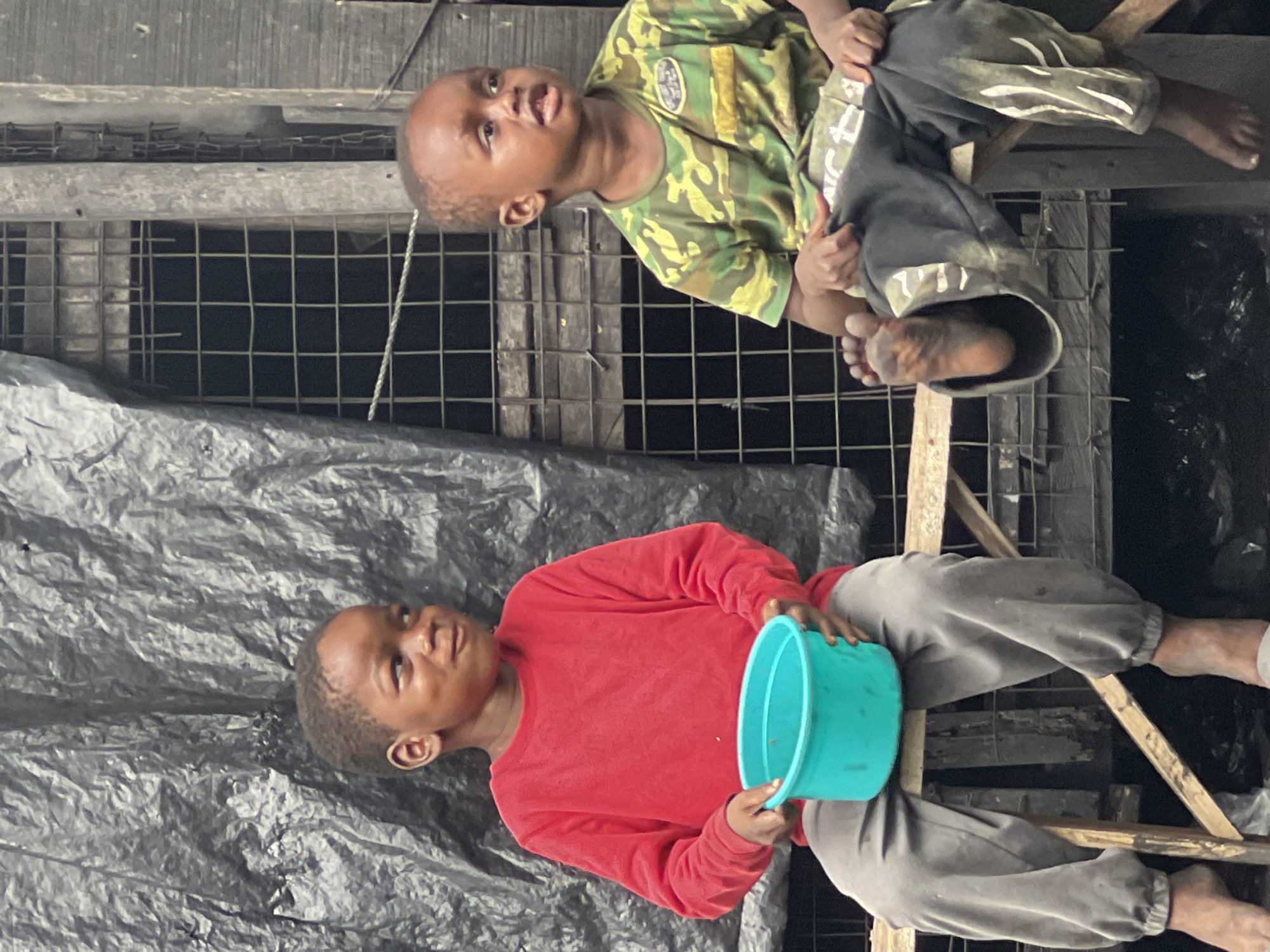 In Nkpor, Nigeria, two young boys are seated on a makeshift wooden bench against a backdrop of a crinkled black tarpaulin. The boy on the left, wearing a red long-sleeved shirt and grey sweatpants, is holding a turquoise bucket in his lap. His eyes are looking to his right. The boy on the right, dressed in a camouflage t-shirt and black sweatpants with white markings, has his right leg extended forward, with his foot resting on the bench. He is looking to his left. Behind them, a metal grate is visible through a gap in the tarpaulin, suggesting a structure with an opening. The overall environment appears to be an outdoor, likely informal, setting.