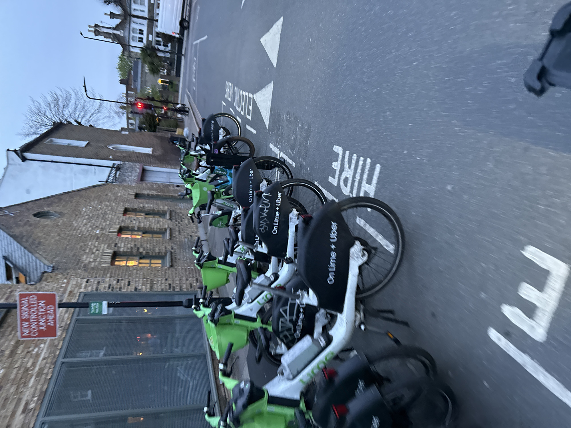 The image shows a row of Lime and Uber electric bicycles parked on a street in London, United Kingdom. The bicycles are white and green and have black seat covers with "On Lime + Uber" printed on them. The street has "HIRE" and "ELECTRIC BIKES" painted on it. There is a road sign that reads "NEW SIGNAL CONTROLLED JUNCTION AHEAD". The background includes buildings, trees, a traffic light, and a car. The photo appears to have been taken at dusk or dawn.