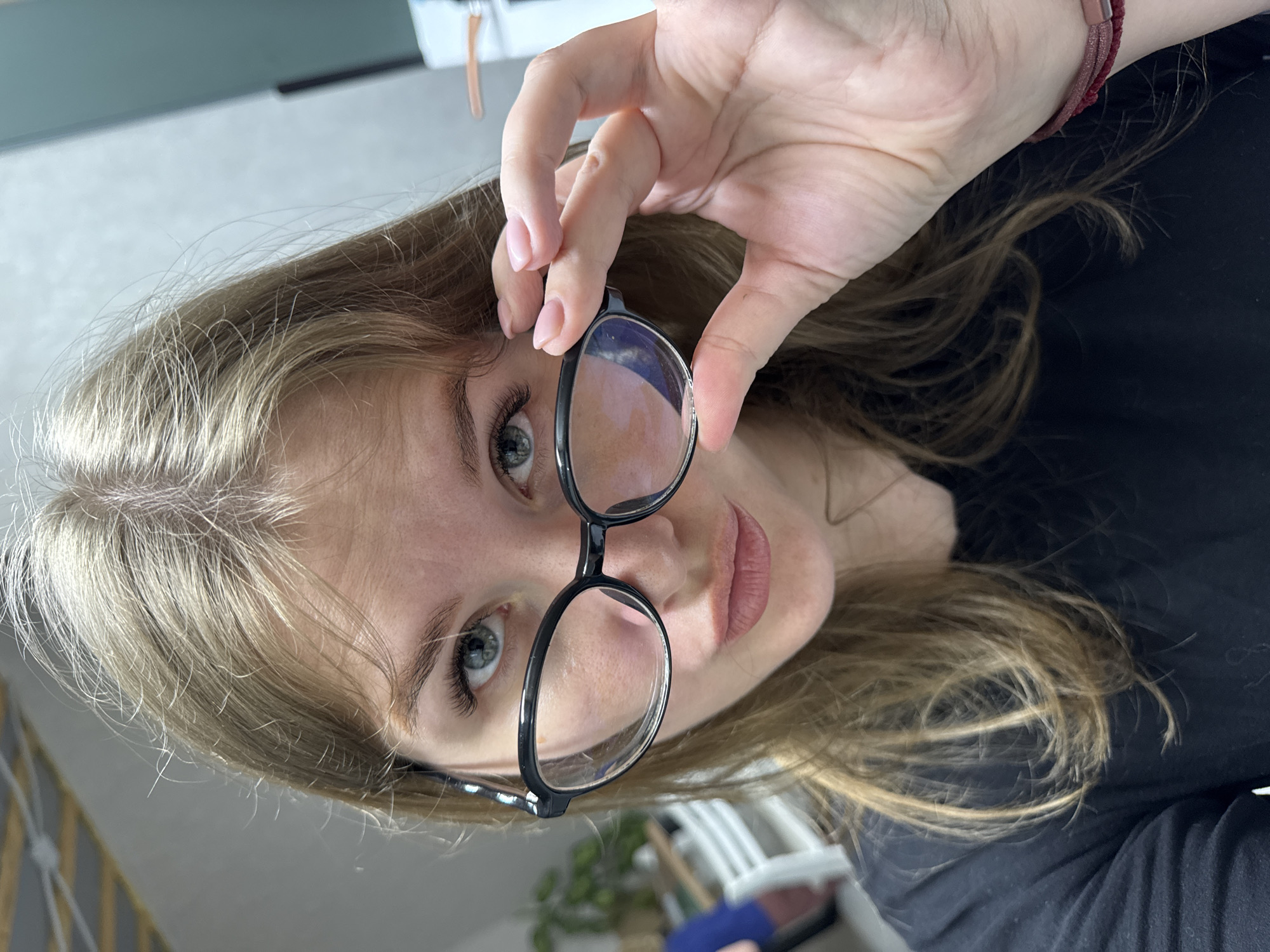 A woman with long blonde hair and light eyes holds a pair of black-rimmed glasses up to her face. Her hand is visible, gripping the glasses frame. She is wearing a dark-colored shirt. The background is blurry, but includes a light-colored wall. The image appears to have been taken indoors, from a close range. The city of Gornyi (Dvurechenskoe S/P), Russia cannot be determined.