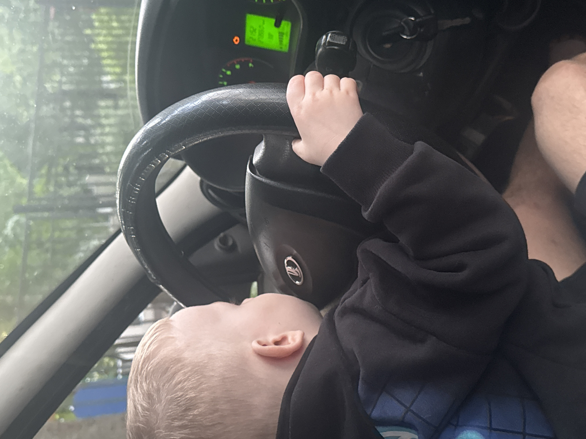 A blonde-haired child is sitting in the driver's seat of a car, holding the steering wheel. The child is wearing a dark hooded sweatshirt. The car's dashboard is visible, with a green digital display showing numbers. The exterior of the car is visible through the windshield, with trees and a blue object in the background. The scene appears to be outdoors, possibly in a residential area of Saint Petersburg, Russia.
