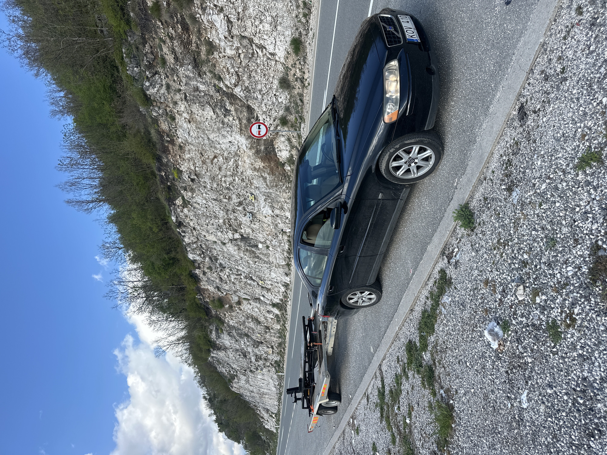 A black Volvo sedan with the license plate KOV 410 is parked on the right shoulder of a two-lane road next to a rocky cliff face. The car is attached to a small, flatbed tow dolly. A round, red and white traffic sign, indicating a prohibition, is posted on the cliff side of the road. The location appears to be outdoors in a mountainous area in Očinići, Montenegro, as evidenced by the landscape. Debris and small vegetation are visible on the gravel shoulder. The sky is partly cloudy.
