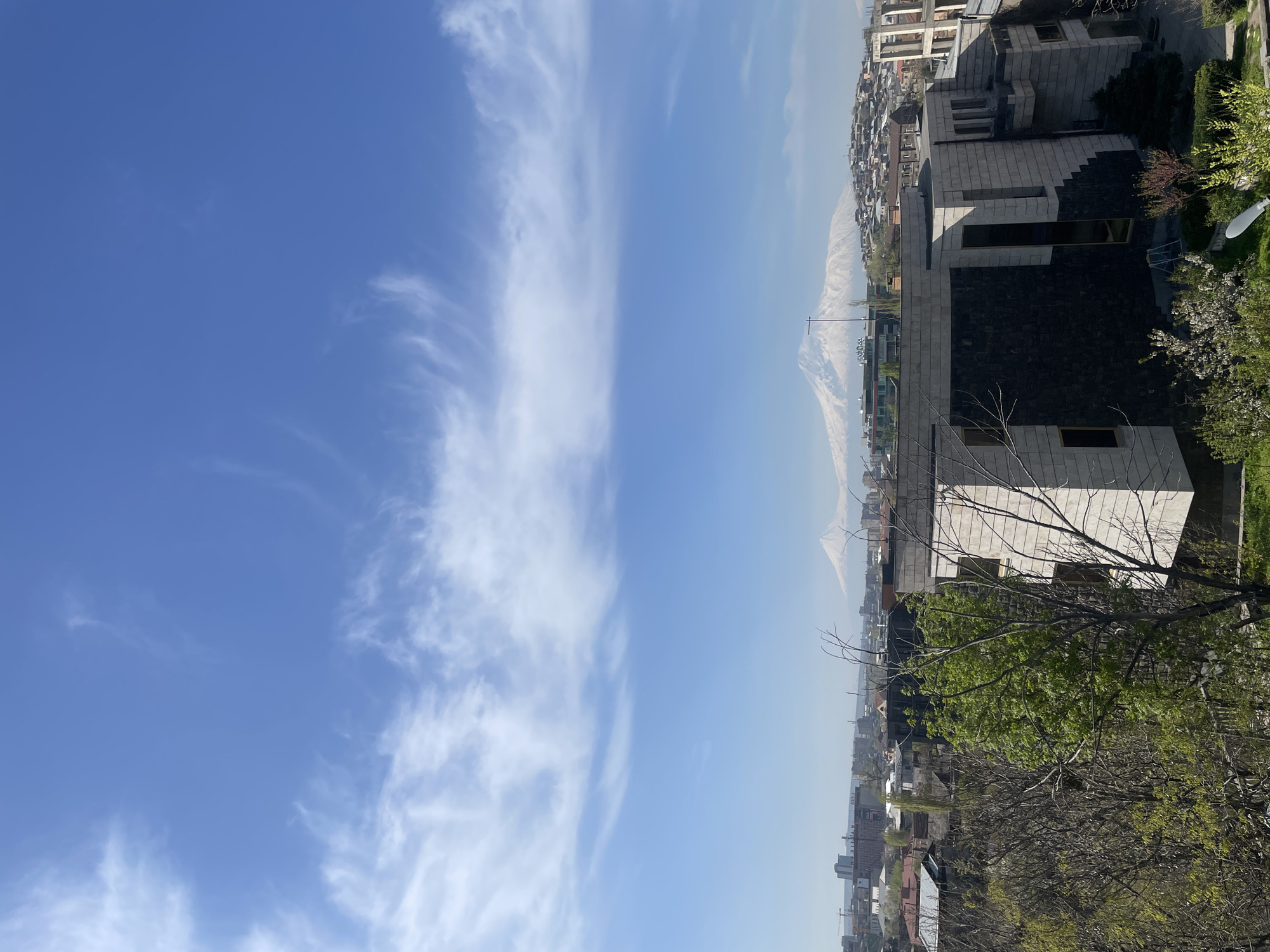 The image captures an expansive daytime view of a city with a majestic snow-capped mountain in the distance, likely Mount Ararat, given the location context of Yerevan, Armenia. The scene is oriented vertically in the provided image, but depicts a horizontal panorama when corrected.
The upper portion of the frame features a bright blue sky adorned with delicate, wispy white clouds, indicative of fair weather. Below the horizon, the sprawling cityscape unfolds under a clear sky, appearing slightly hazy in the distance. The prominent mountain, its upper slopes covered in snow, dominates the background, rising above the urban landscape.
In the mid-ground, numerous buildings of varying sizes and architectural styles populate the city, extending towards the base of the distant mountain. A notable modern building with gray and dark stone facades, featuring large rectangular windows, stands out in the foreground. Below this building, a vibrant green tree with fresh spring foliage occupies the lower right, partially obscuring other structures. To the left and slightly further back, other trees with bare branches are visible, suggesting early spring. A white, dish-like object is partially visible next to the prominent building on the right.
The image suggests a bright, sunny day, likely late morning or early afternoon, with good visibility. No people or visible text are present in the photograph.