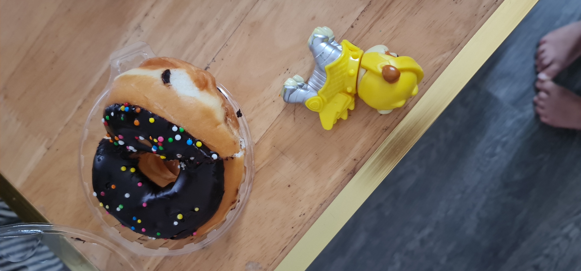 A plastic container of two donuts, one glazed and one chocolate with sprinkles, sits on a wooden surface next to a yellow and silver toy figure resembling a dog knight. Part of a human foot is visible in the background, which appears to be a section of flooring bordering the wooden surface. The photo does not provide enough information to determine a location in Đà Nẵng, Vietnam.