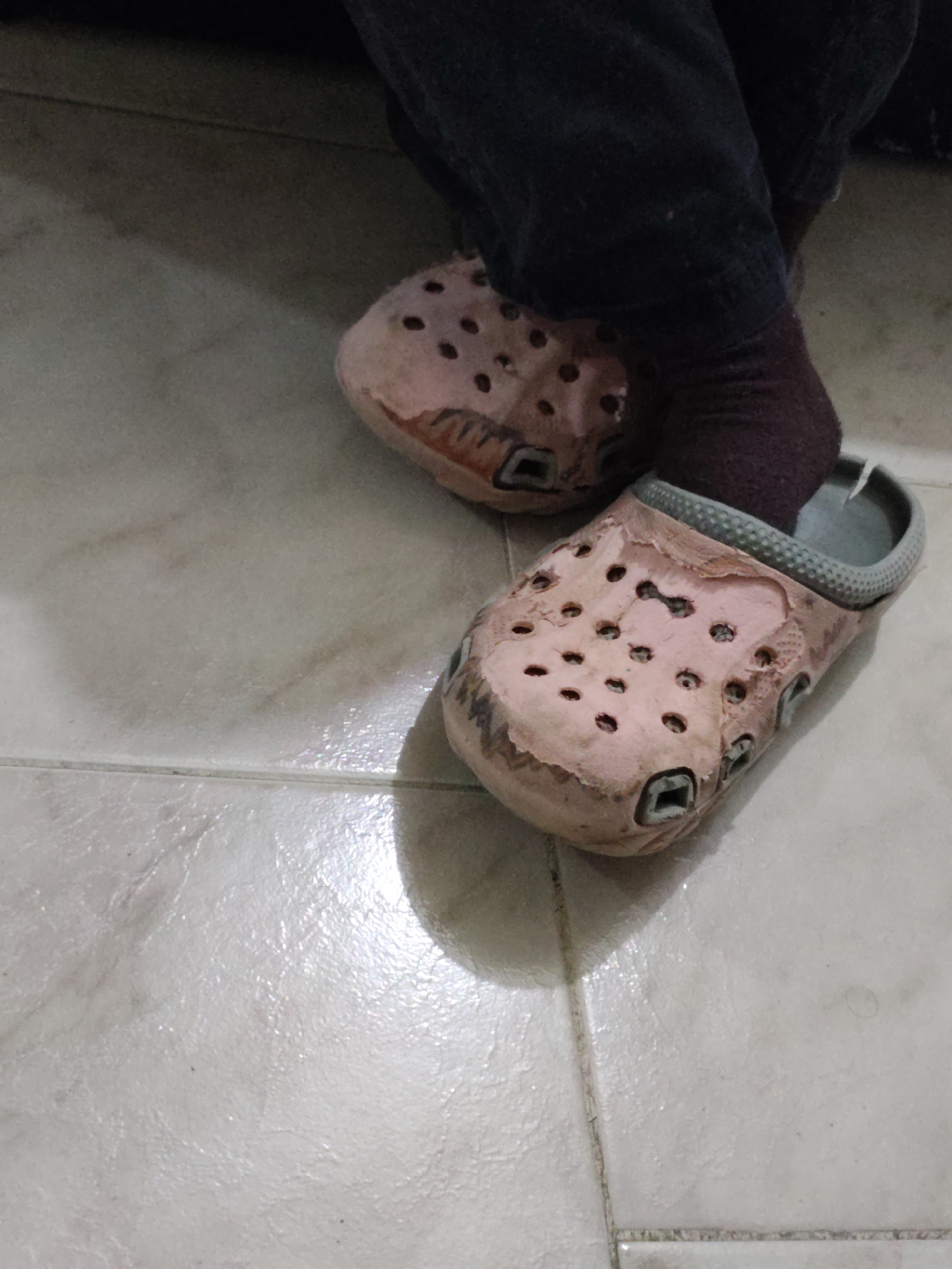 A pair of worn, light pink Crocs are shown from a high-angle shot, placed on a tiled floor. The Crocs have visible scuff marks and wear, particularly around the edges and toe area, revealing darker material underneath. The interior of the Crocs appears to be a light gray. Dark purple socks are visible peeking out from the top of the Crocs. The tiled floor is light-colored with visible grout lines and a slight sheen, suggesting it may be slightly damp or reflective. The background is dark and out of focus, with hints of dark fabric that may be clothing. The scene appears to be an indoor environment. The image is located in Piloncito, Venezuela.