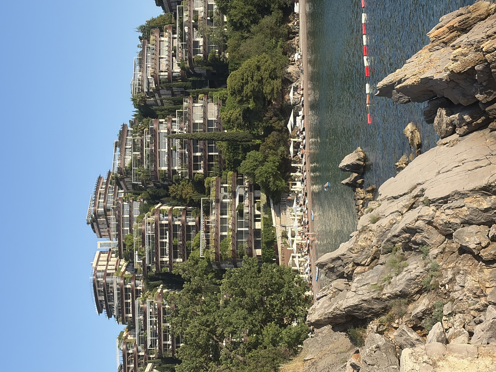 The image depicts a coastal scene in Budva, Montenegro. In the background, a terraced building complex with balconies is visible, nestled among green trees. The buildings have a modern architectural style. In the mid-ground, a beach area is populated with sunbathers and beach umbrellas. A body of water, possibly the Adriatic Sea, is present, with a red and white safety buoy line visible. The foreground features rocky terrain, with large gray rocks jutting out, suggesting a cliffside view. The sky is clear and blue.