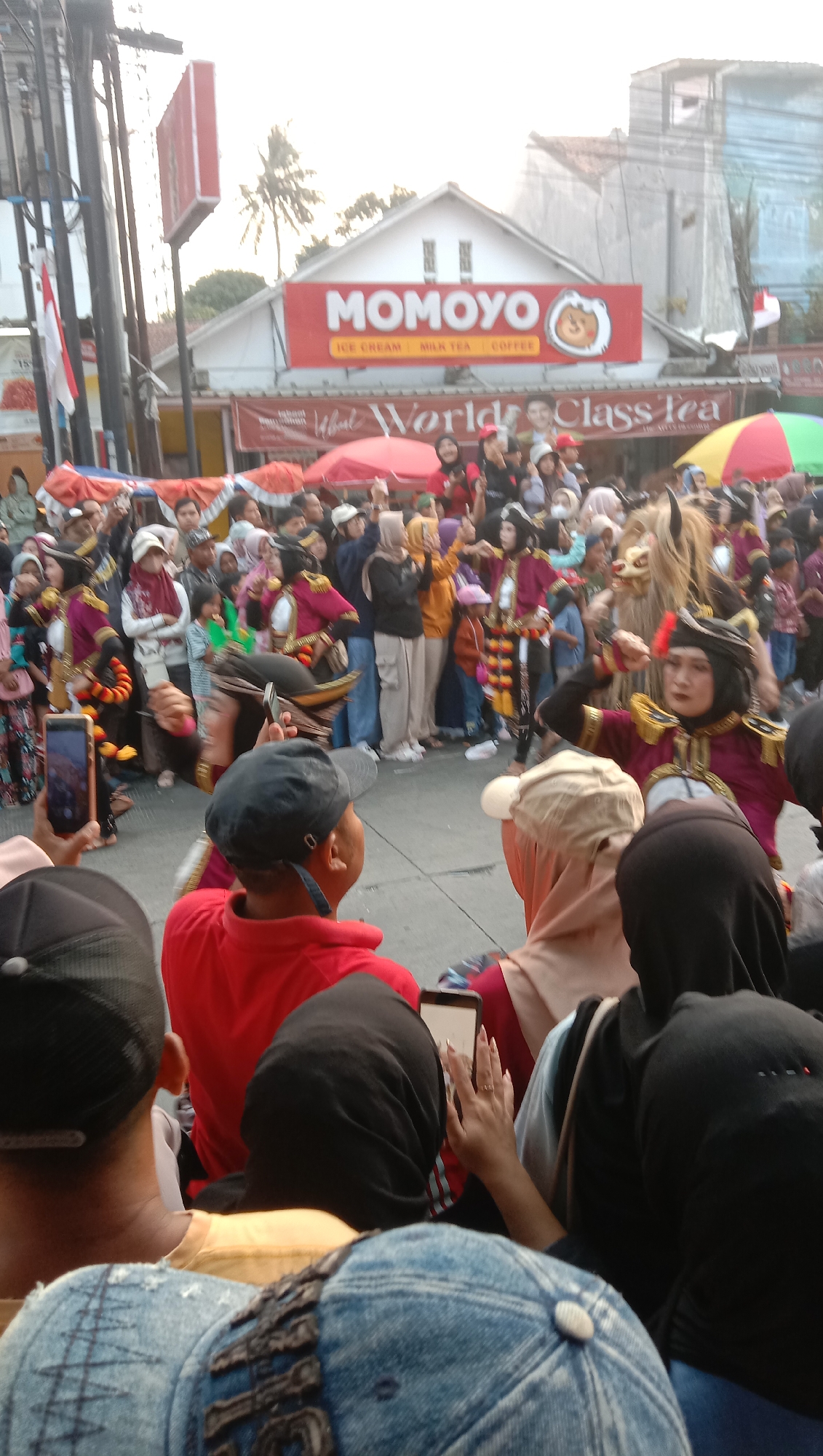 The image depicts a lively outdoor scene, likely a street event or festival, in Jatisawit, Indonesia. The focal point is a procession or performance with individuals dressed in colorful, traditional costumes, some with elaborate headdresses and makeup. One performer appears to be wearing a mask or costume resembling an animal. A crowd of onlookers lines the street, many holding up their mobile phones to capture the event. In the background, a building displays a prominent red sign with the text "MOMOYO" and smaller text below reading "ICE CREAM MILK TEA COFFEE". Further signage indicates "World Class Tea". The atmosphere is one of celebration and public engagement, with a mix of cultural elements and modern technology present.