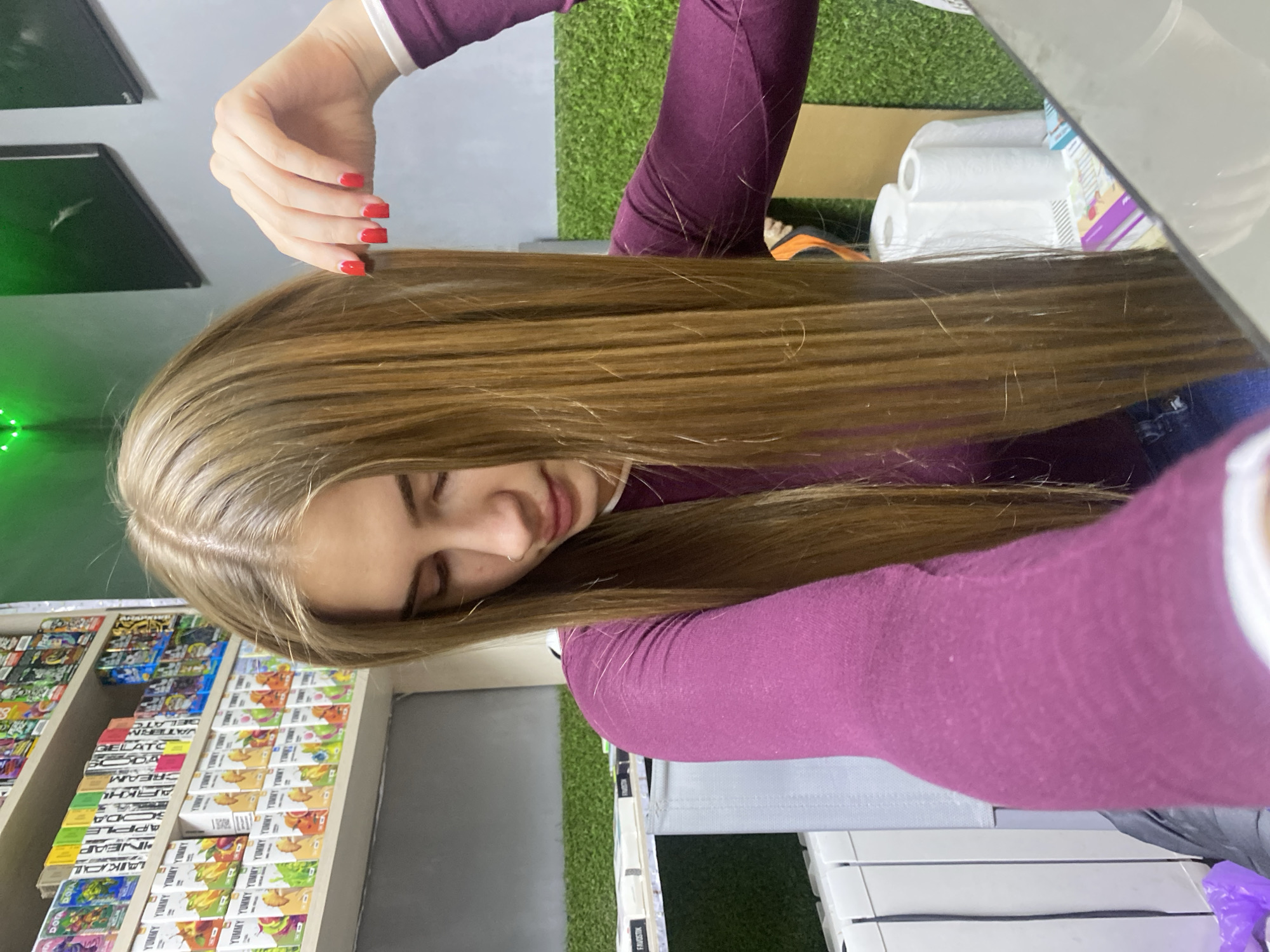 The image shows a young Caucasian woman with long, straight blonde hair and red nail polish. She is wearing a long-sleeved purple top and blue jeans. The woman is looking down and to her left. She is indoors, in what appears to be a shop or retail environment. In the background, there are shelves stocked with various products, possibly vape products or e-liquids, as indicated by the colorful packaging. There is also a display of green artificial turf and some rolls of paper. The lighting is bright, and the overall scene is well-lit. The location is noted as Дьяково, Russia.