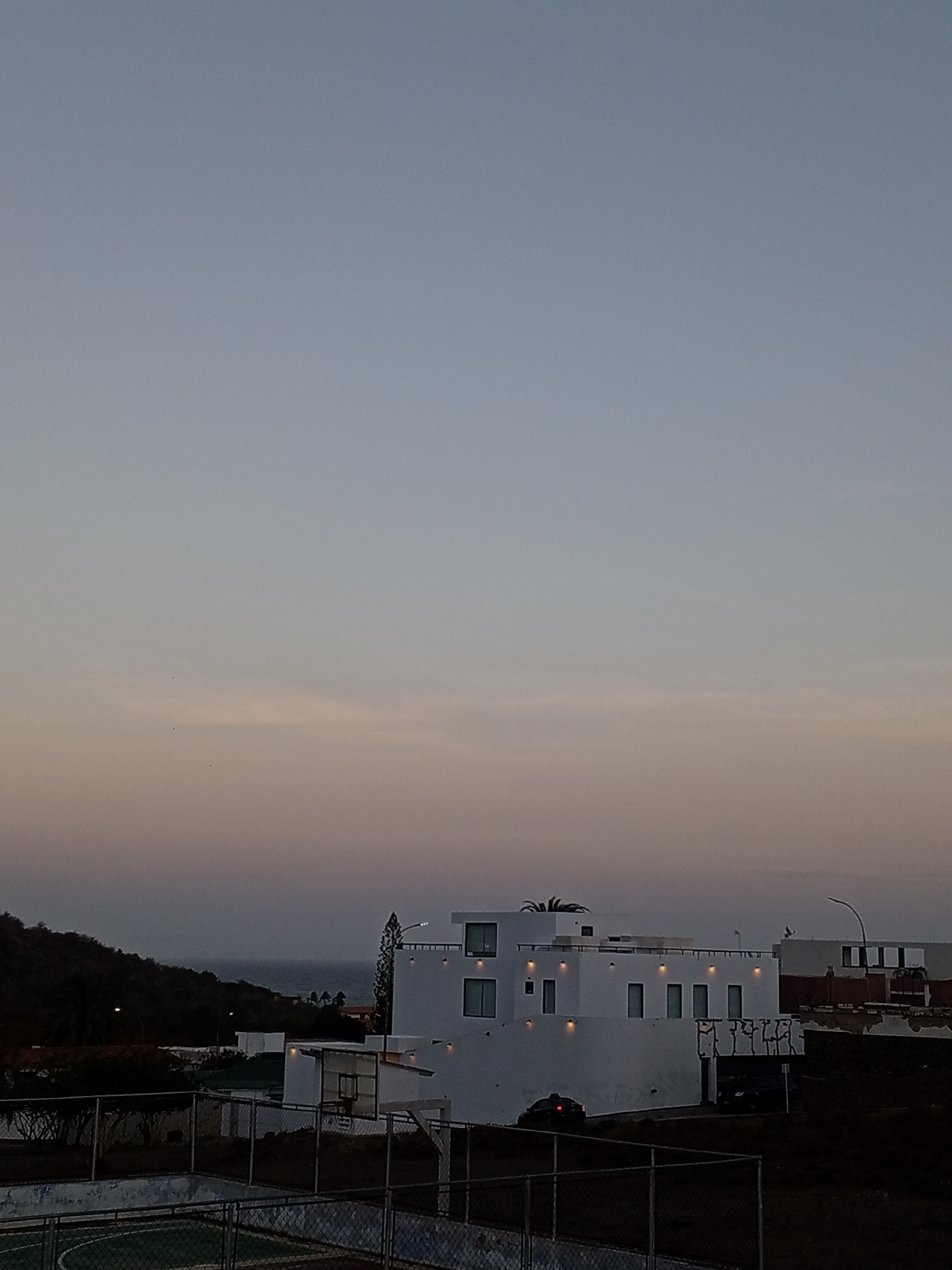 The image depicts a twilight scene in Pampatar, Venezuela. In the foreground, a fenced basketball court is visible. Beyond the fence, a two-story modern white building with rectangular windows is the focal point. To the left of the building, a tree-covered hillside slopes down to the ocean, partially obscured by a layer of haze. To the right, parts of nearby houses are present. The sky is a gradient of blues and oranges.