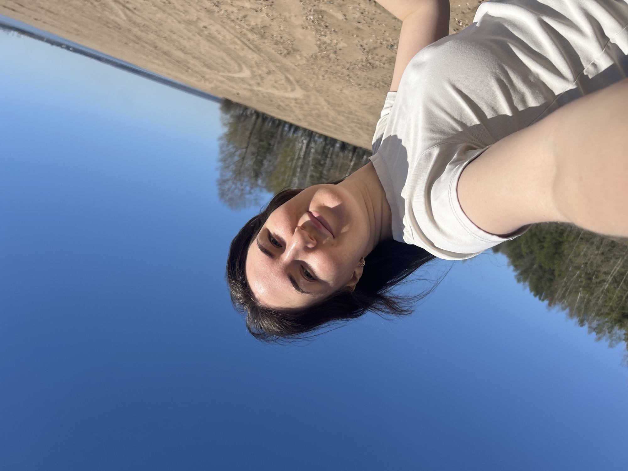 A woman is pictured from a low angle. The woman is lying on a beach in Noviy, Russia, and is wearing a white t-shirt. The background of the image shows a blue sky, sand, trees, and a glimpse of water. The woman's face is visible, and she appears to be looking at the camera. The scene is outdoor during the day.
