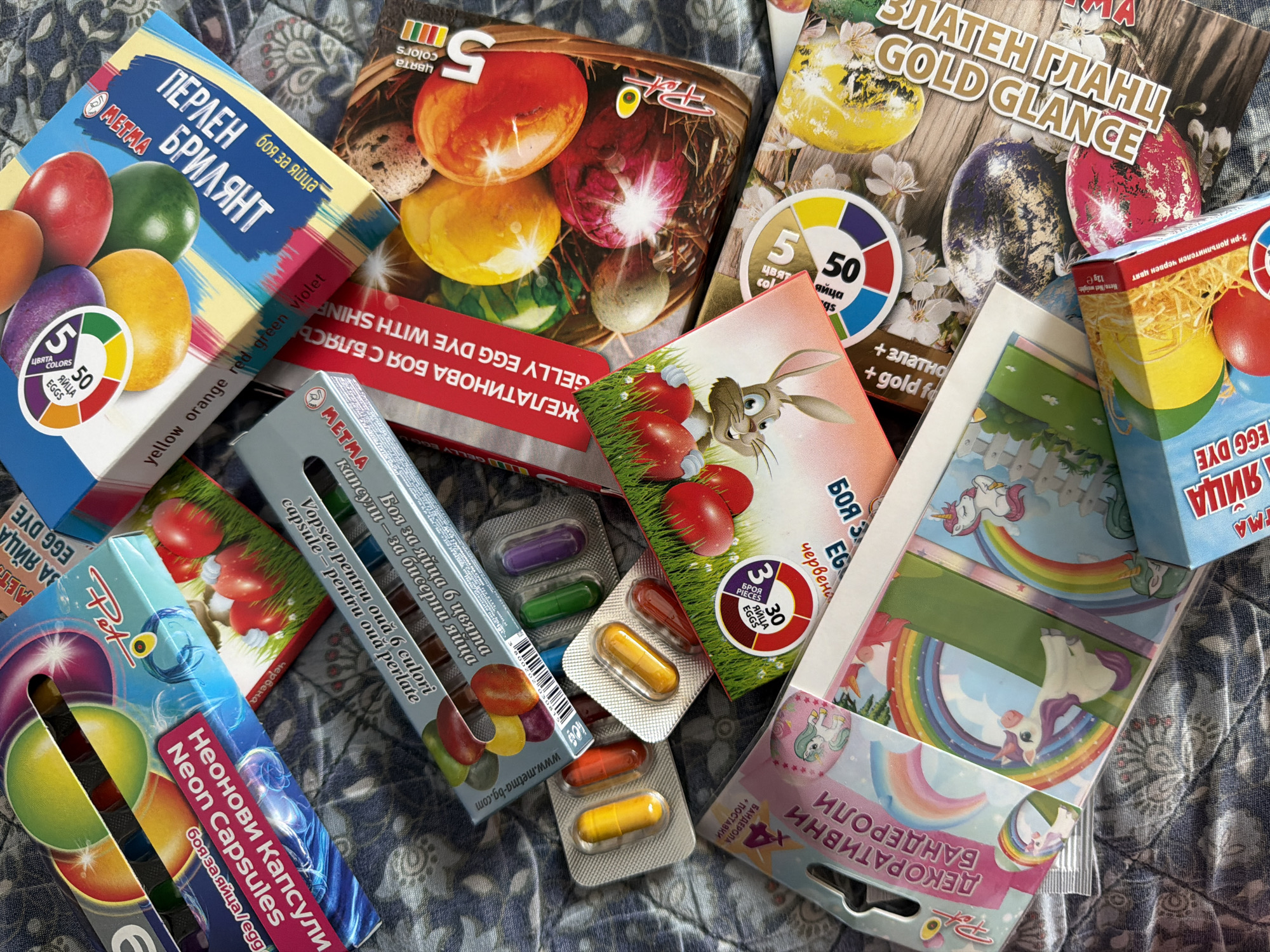 The image shows a collection of Bulgarian Easter egg dyeing and decorating kits scattered on a patterned surface, possibly a bedspread. The kits are produced by the company "Metma". The kits include a variety of methods for coloring and decorating eggs:
* Capsules of egg dye, some with "Neon" colors
* Powdered egg dye
* "Gold Glace" egg dye
* Egg wraps with unicorn themes.
The brands of the kits, such as “Pot" are also displayed. There are loose dye capsules on the surface. The text on the boxes is in Bulgarian and English. There is no indication of the location of the picture being Pazardzhik, Bulgaria.
