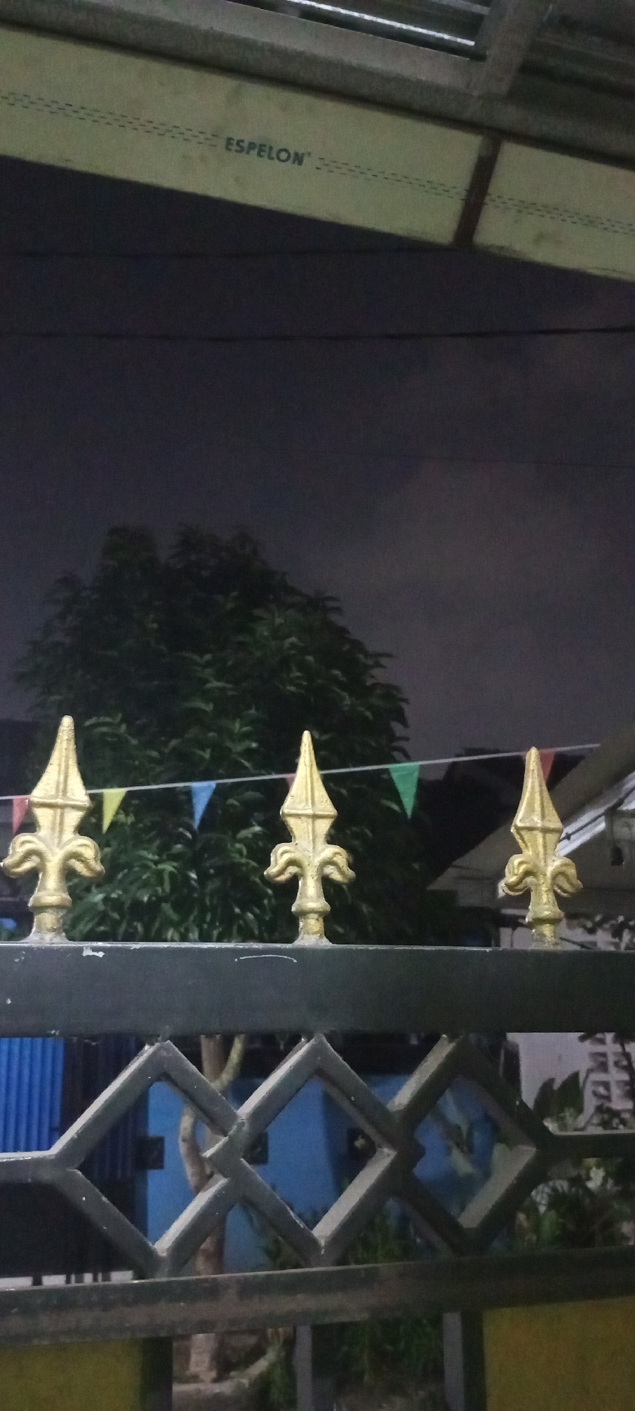 The image shows a nighttime view of a fence with decorative gold finials. Behind the fence, a tree with dark green foliage is visible against a dark, cloudy sky. A string of triangular flags in various colors is draped across the scene above the fence and in front of the tree. The fence itself is black and features a diamond pattern. In the background, a blue wall and some greenery can be seen. The text "ESPELON" is visible on a light-colored surface in the upper portion of the image. The setting appears to be residential, possibly in Lubangbuaya, Indonesia, given the context.