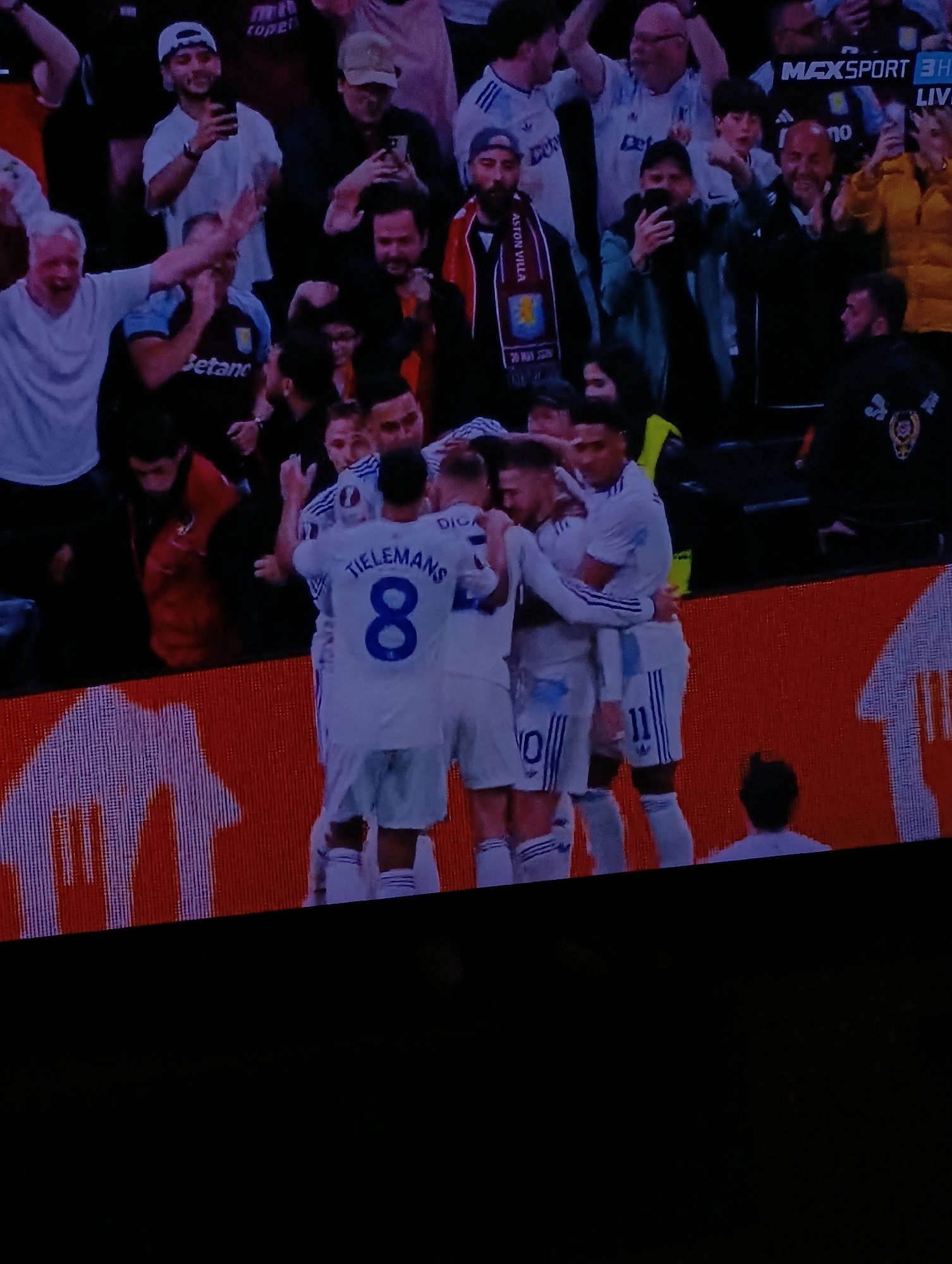 The image captures a scene from a football match, likely broadcast on Max Sport, a sports channel (as indicated by the "MAXSPORT 3H LIVE" text in the upper right corner). A group of players in white jerseys are huddled together, celebrating. One player has "TIELEMANS 8" on the back of his jersey, while another has "11" on his shorts. The word "DIGA" is visible as well. Spectators are visible in the background. Some are recording with their phones, while others are celebrating or watching the game. A banner or scarf with "ASTON VILLA" is visible in the crowd. The football pitch appears to be surrounded by a brick-like wall. It is not possible to confirm the location in the city of Plovdiv, Bulgaria, only based on this single image.