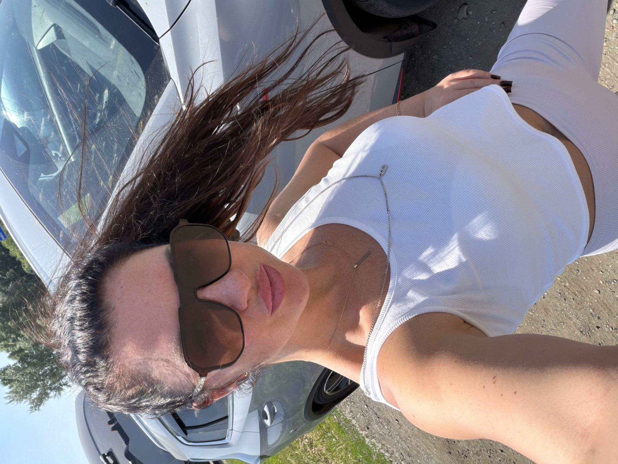 A woman is captured in a selfie outdoors, with a grey SUV partially visible in the background. She is wearing a white cropped tank top and light-colored leggings. Her dark hair is pulled back in a ponytail and is being blown by the wind. She has large sunglasses on and is wearing a delicate silver necklace. The ground appears to be gravel or dirt, suggesting a rural or suburban setting. The sky is clear and bright, indicating a sunny day. No specific landmarks of Пригородный, Russia are identifiable in the image.
