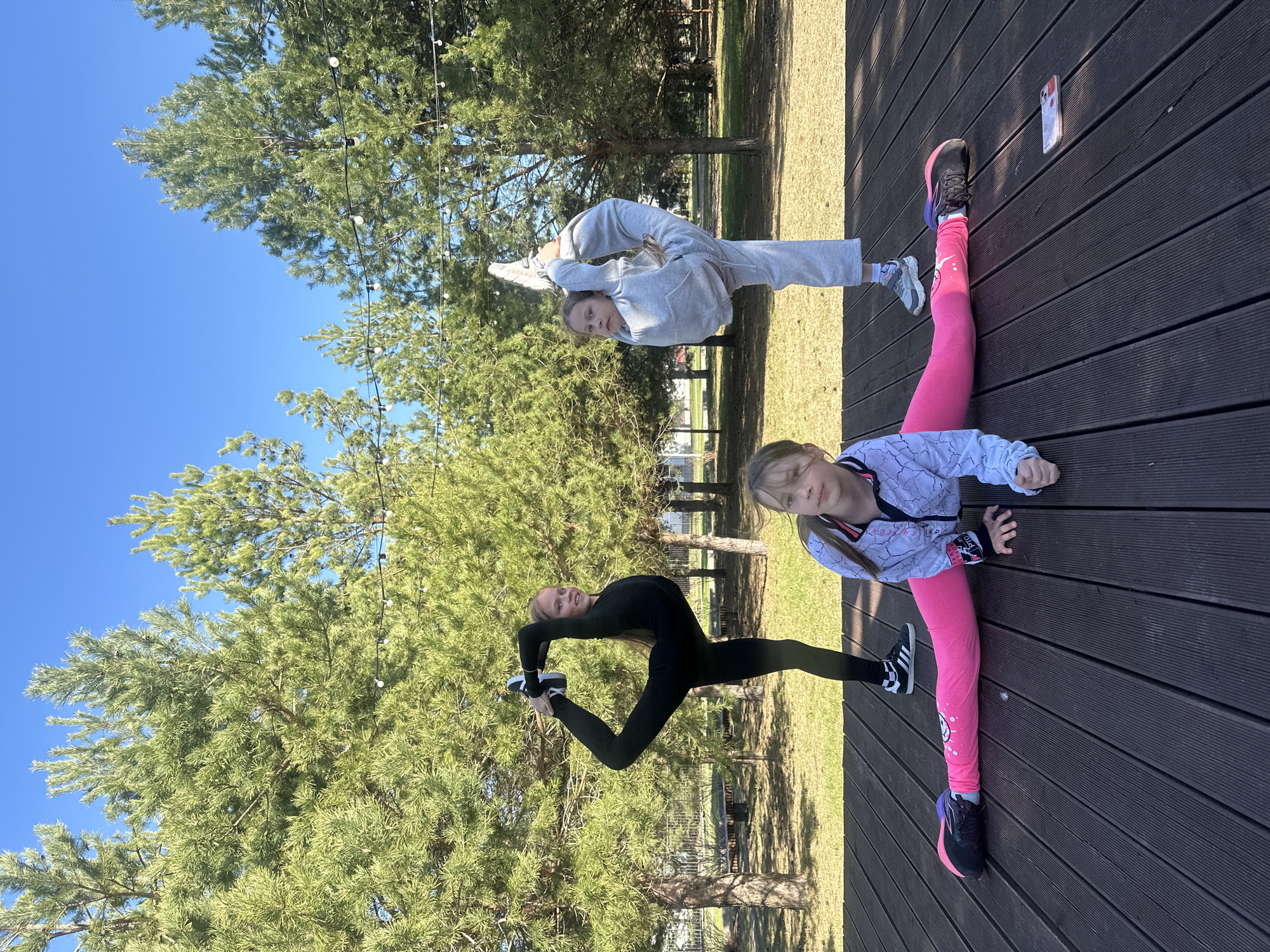Two young girls are performing acrobatic stretches on a dark brown wooden surface, possibly a stage or patio, outdoors. The girl closer to the viewer wears pink leggings, a white jacket with black and pink details, and black sneakers with pink and blue soles. She is doing a split. The second girl, positioned higher in the frame, wears a gray tracksuit and white sneakers, attempting a backbend while holding her feet. Behind them are pine trees and a bright blue sky. The location appears to be a park in Русский Вожой, Russia, based on the provided context. A mobile phone lies on the wooden surface near the first girl's foot. Another girl wearing a black jumpsuit and black and white sneakers does a similar stretch pose like the first girl.