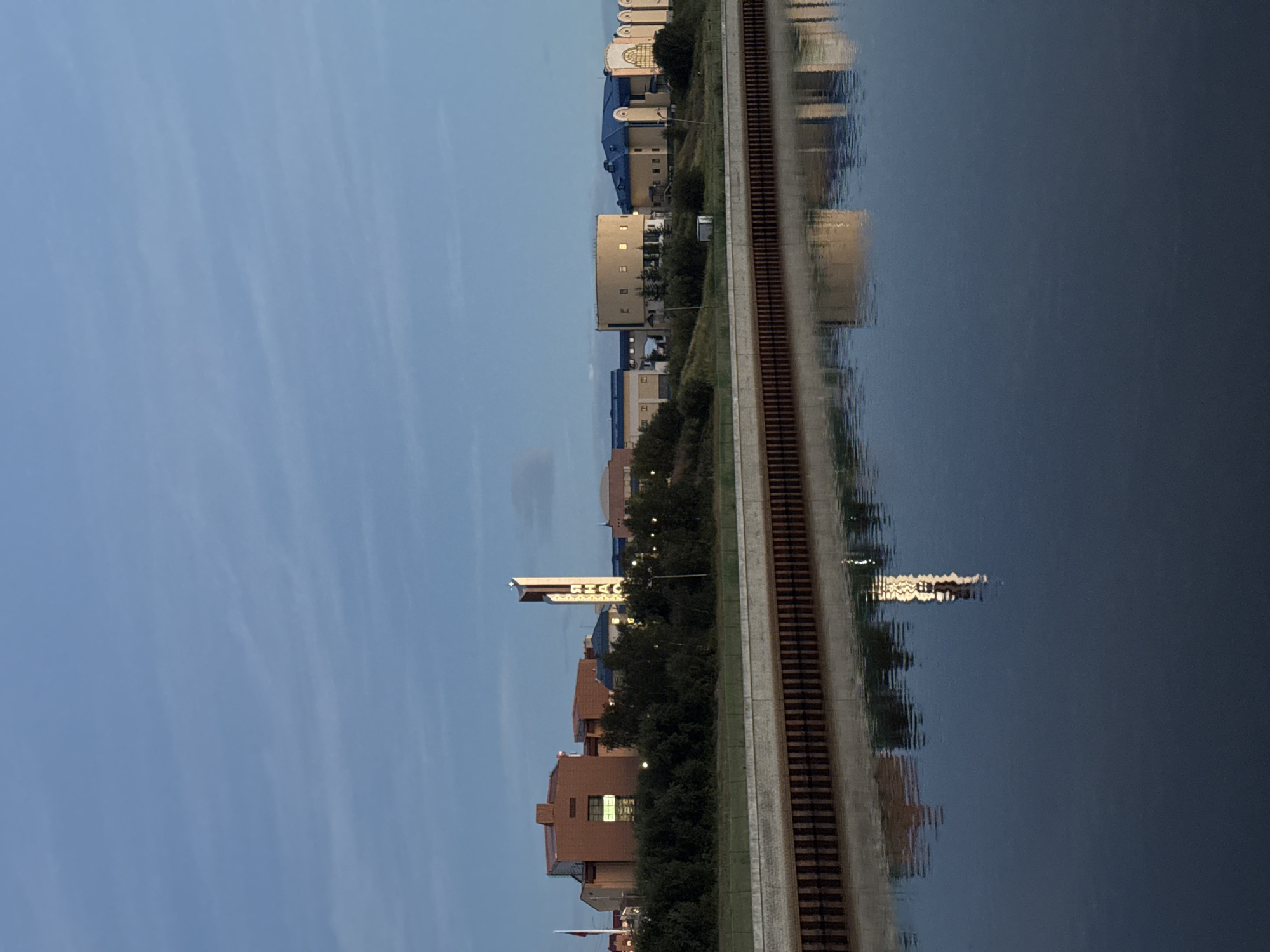 The image shows a wide, calm river with reflections of buildings and a tower on the opposite bank. The buildings are of various architectural styles, including a prominent reddish-brown brick structure, a cylindrical building with yellow walls and round windows, and other buildings with blue roofs. A tall, slender tower with illuminated vertical lettering is visible, which is part of the "Salekhard" sign, a well-known landmark in the city. The riverbank is lined with a wooden retaining wall. The sky is a pale blue with faint clouds, suggesting it is either early morning or late evening. Lush green trees and vegetation grow along the elevated bank behind the buildings. No people are visible in the image. The scene depicts a quiet, urban waterfront in Salekhard, Russia.