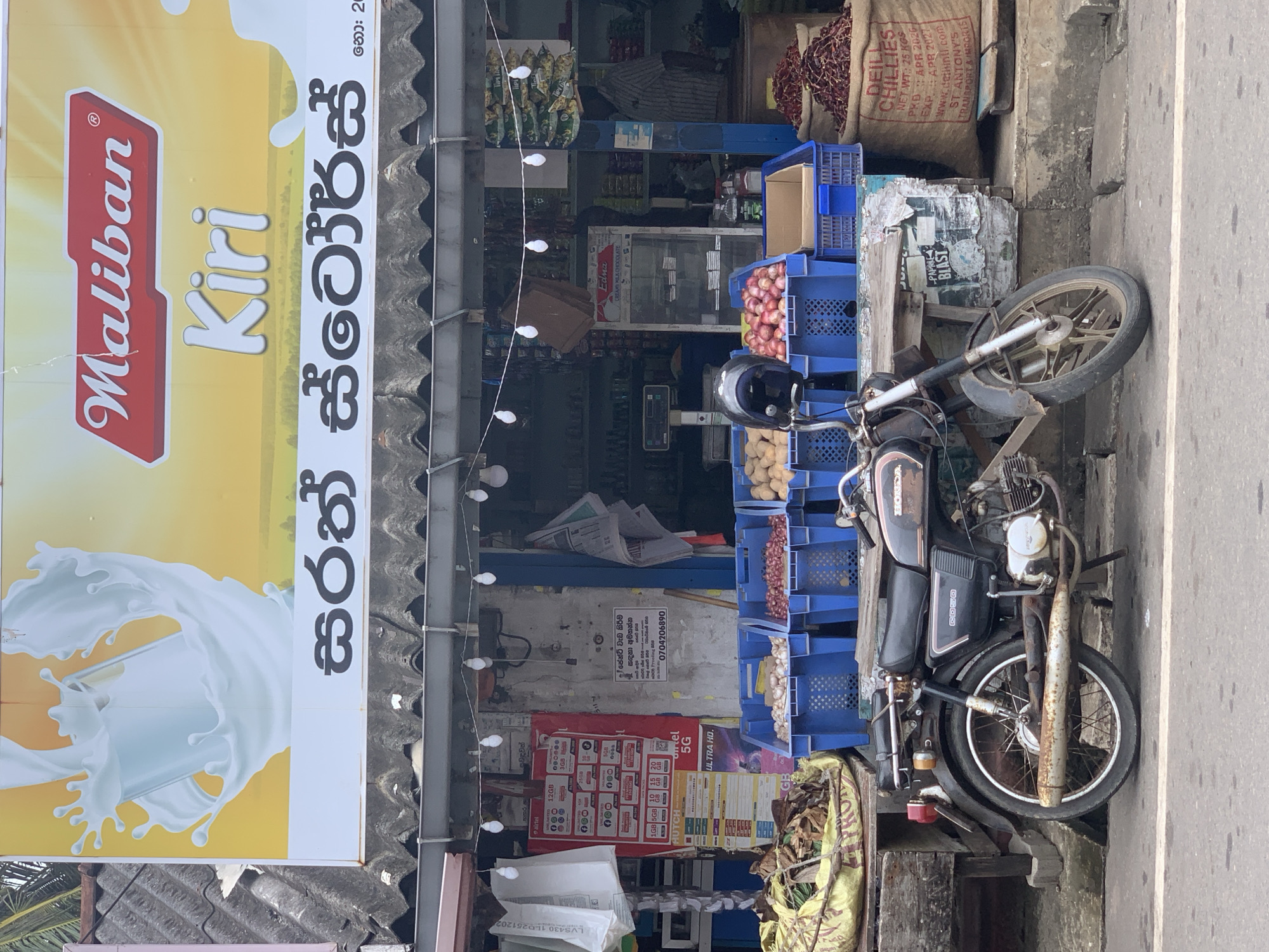 This is a medium shot of a roadside shop in Ahangama, Sri Lanka. A vintage, dark-colored motorcycle with significant rust is parked in front of the shop. The shop has a prominent yellow and white awning with the Maliban Kiri logo and text in Sinhala script. Underneath the awning, the shop displays an assortment of goods, including blue crates filled with onions, potatoes, and red apples. Bags of dried chilies and other produce are also visible. String lights with visible bulbs are hung across the storefront. The overall scene depicts a typical Sri Lankan market environment.
