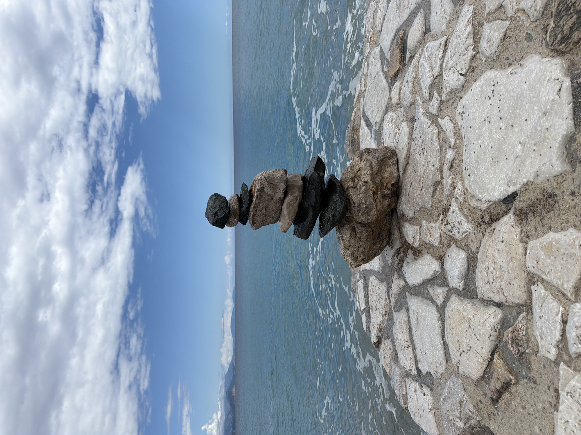 The image displays a serene coastal scene in Dubraljevina, Montenegro, under a bright blue sky with scattered white clouds. A stack of approximately six to seven rocks, forming a cairn, is prominently featured on the right side, balanced on an irregularly paved stone surface at the water's edge. The rocks in the cairn vary in color from dark grey and black to lighter browns and greys.
The stone pavement, made of light-colored, angular stones set in mortar, forms a barrier or path along the sea. To the left of this stone edge, the blue-green sea extends to the horizon. Gentle waves are visible, breaking against the paved surface and creating white foam. In the distance, faint outlines of mountains or landmasses can be seen across the water, beneath the clear sky. The sky itself is adorned with fluffy white clouds, more concentrated on the left side of the frame. The overall impression is one of tranquility and natural beauty during a clear day. No people or visible text are present. The rock cairn suggests human presence and activity, possibly for meditation or as an artistic marker.