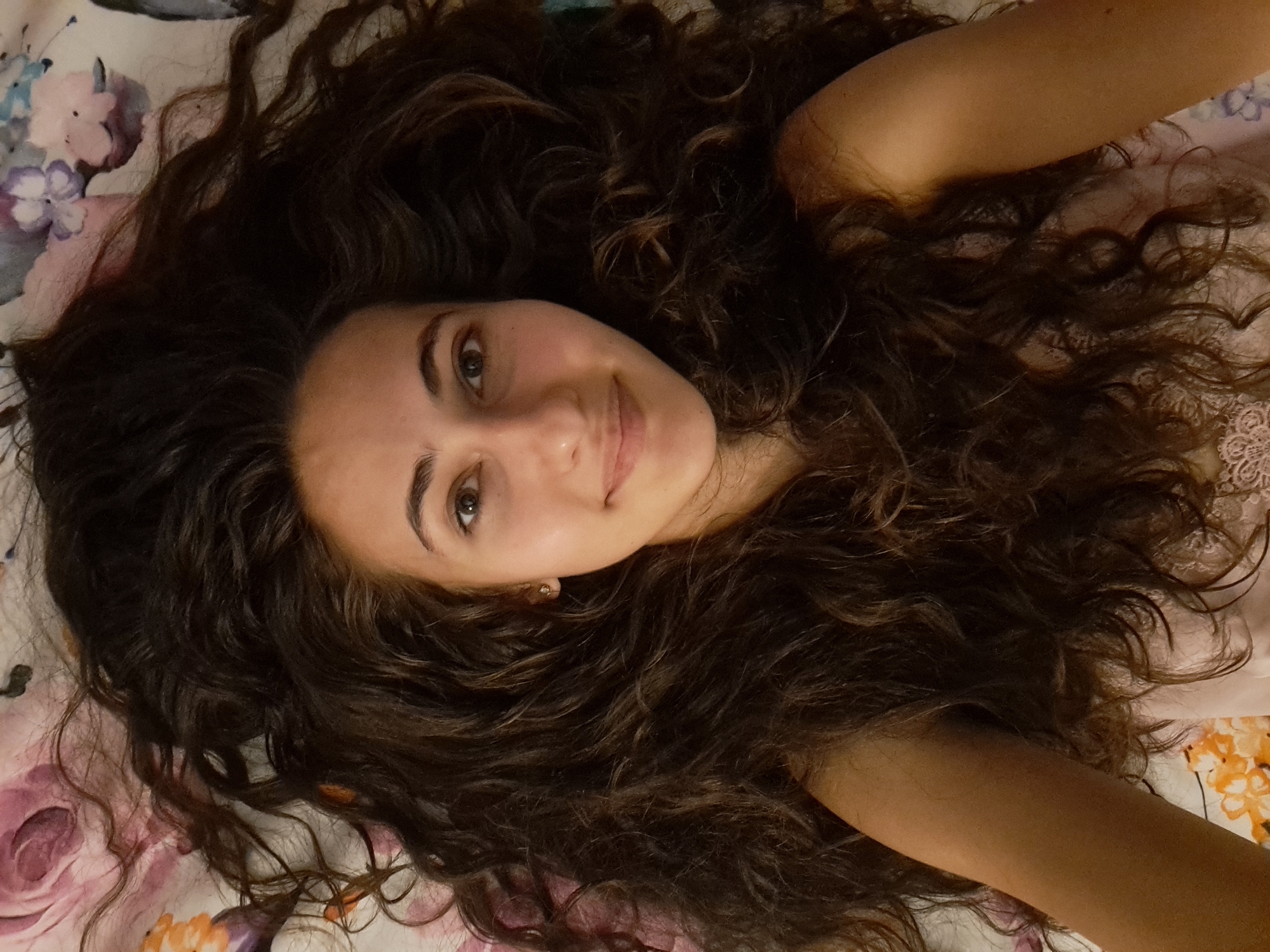 A young woman with long, dark, curly hair is lying on a floral-patterned surface, likely a bed or blanket. She is looking up and slightly towards the camera, with a gentle smile. Her hair is spread out around her head, filling a significant portion of the frame. She is wearing a light-colored top with lace trim. The lighting suggests an indoor setting. The background features a floral pattern with various colors, including pink, purple, orange, and white.