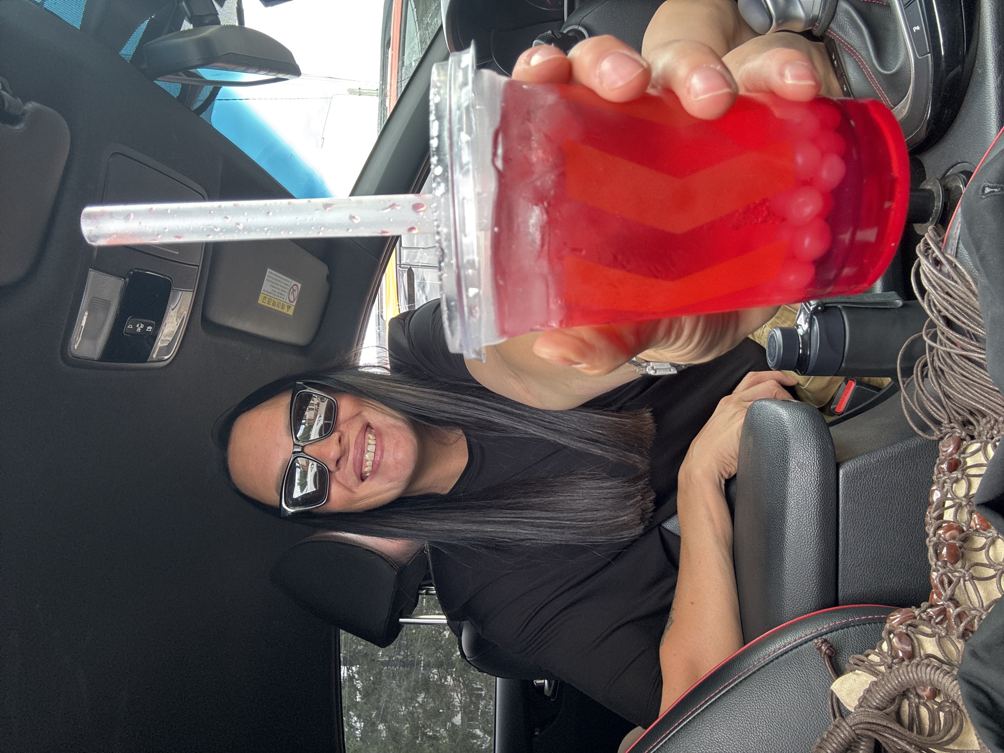 A woman with long, dark hair and sunglasses is sitting in the driver's seat of a car, holding a clear plastic cup filled with a red beverage and what appear to be boba pearls. The cup has a clear plastic lid with a white straw. The woman is smiling and looking at the camera. The car's interior is visible, including the dashboard, center console, and a portion of the passenger seat. The background shows trees and a building, suggesting an outdoor location, possibly in a city. The image is a candid shot, likely taken by the woman herself or someone in the car with her. The scene depicts a casual moment, possibly a quick stop for a drink while on the go.