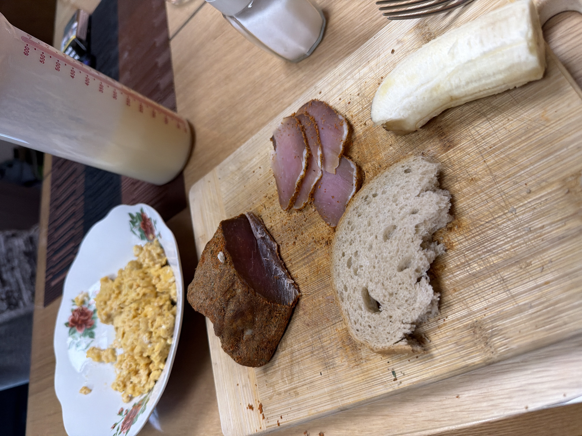The image presents a tabletop scene, likely indoors. A wooden cutting board holds a partially peeled banana, a slice of bread with a bite taken out of it, and two cuts of cured meat, one in slices. To the left, a floral-patterned plate contains scrambled eggs. A measuring pitcher filled with a light-colored liquid and a salt shaker are also visible. A fork is also visible on the cutting board. The location in the city of Panagyurishte, Bulgaria, is not discernable from the provided visual information. There are no people visible.