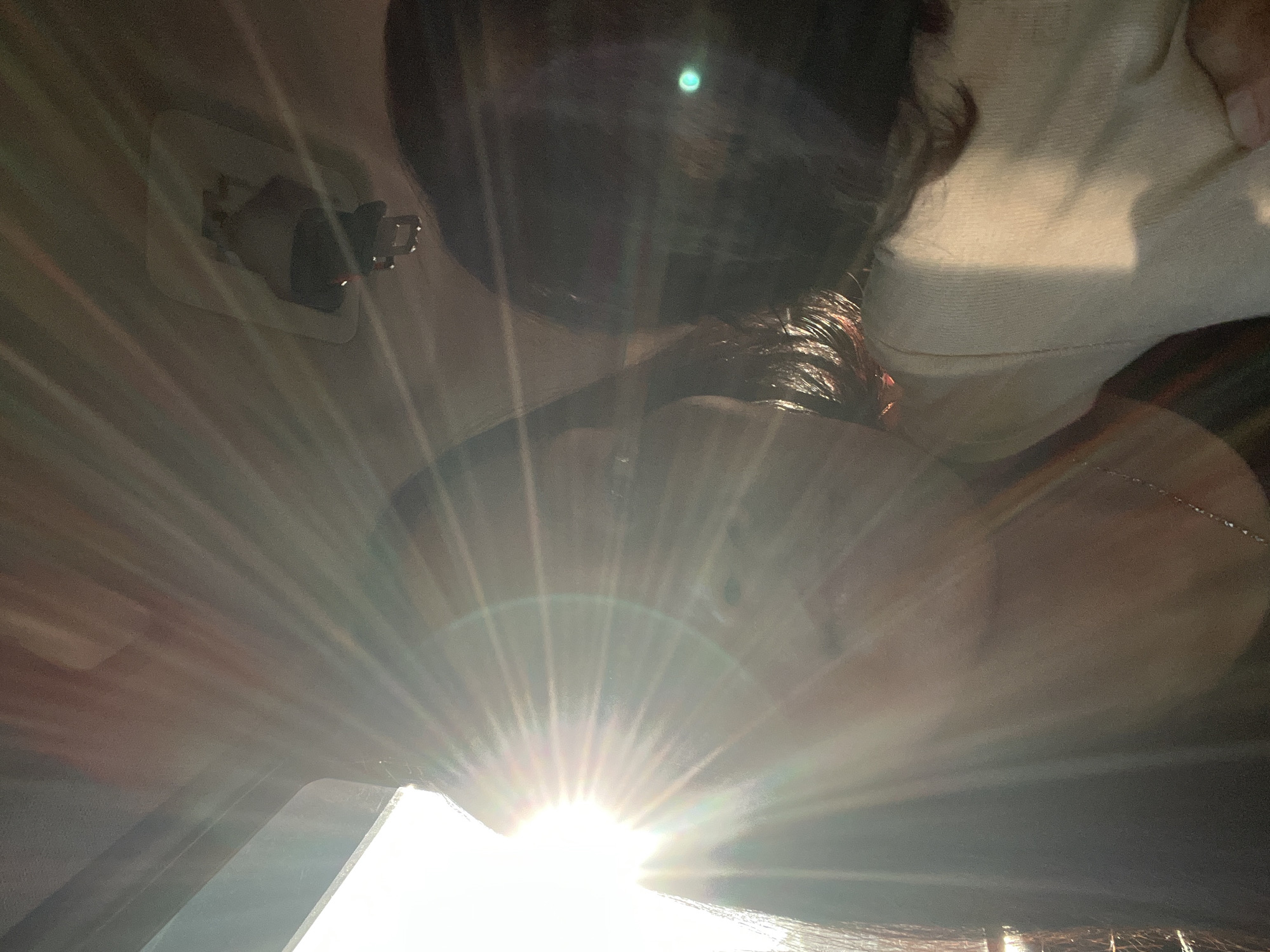 The image depicts a close-up, low-angle shot of a person's face and upper body, with a strong sun flare obscuring much of the detail. The person appears to be lying down or leaning back, with their head tilted upwards towards the bright light. Their dark hair is visible, and a portion of their face is illuminated by the sun. They are wearing a light-colored, textured top.
The setting appears to be indoors, possibly a car or a room with a window. The glare suggests it is daytime, and the sun is quite intense, indicating it might be midday or late afternoon. The sun flare creates a starburst effect with prominent rays extending outwards, originating from a bright, white light source at the bottom of the frame. The background is dark and indistinct, with hints of wooden surfaces and possibly some metallic elements. There are no discernible text or specific location cues visible in the image, other than the provided location context of Getas, Indonesia. The overall mood is somewhat abstract due to the overwhelming light and limited visibility of the subject.
