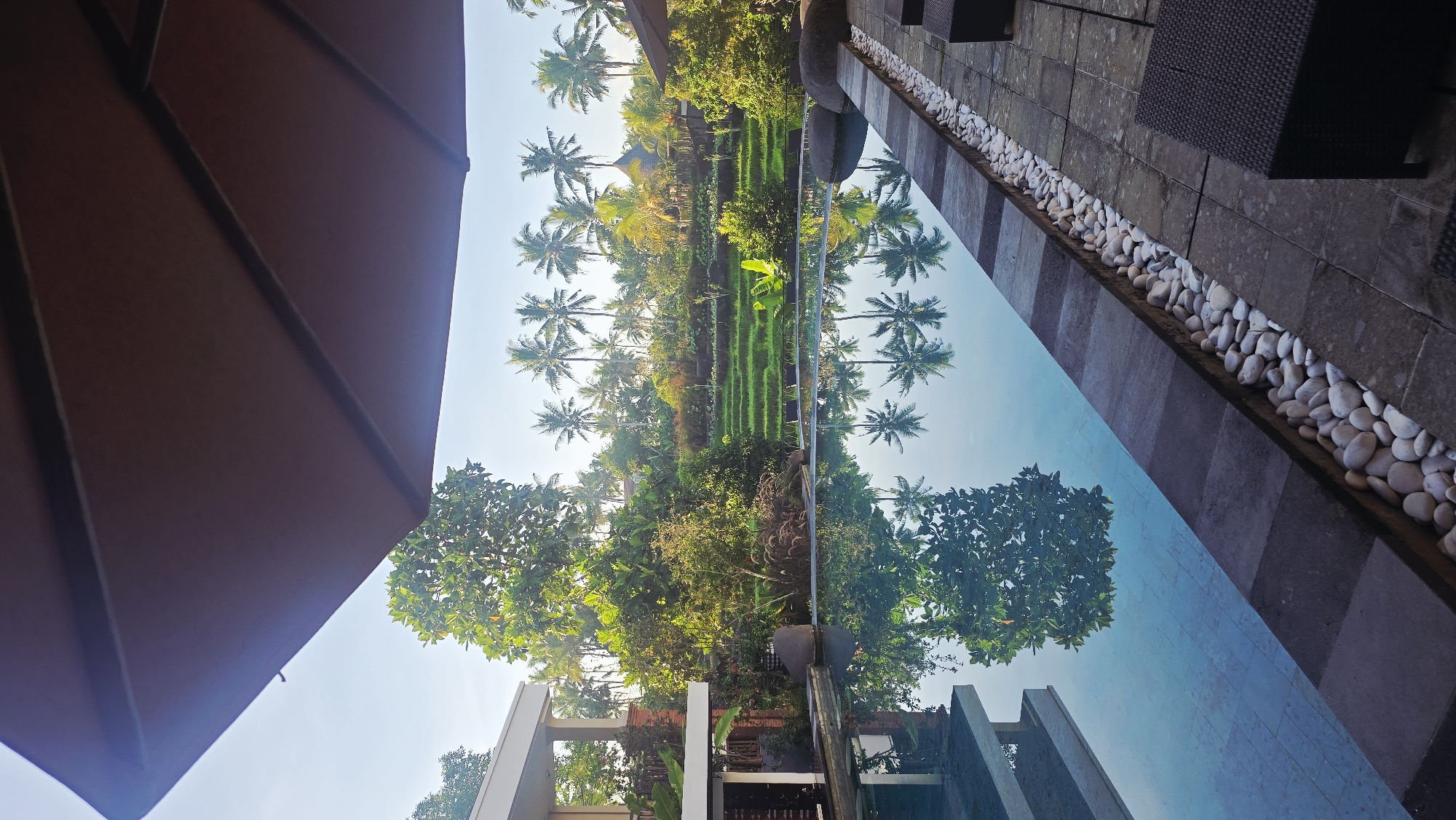The image depicts a reflective pool in Ubud District, Indonesia, mirroring a landscape of rice paddies and palm trees under a bright sky. An umbrella partially obscures the view on the left. The pool's edge is lined with white pebbles and gray stone tiles. Wicker furniture is partially visible to the upper right. The reflection in the pool includes trees and building structures.