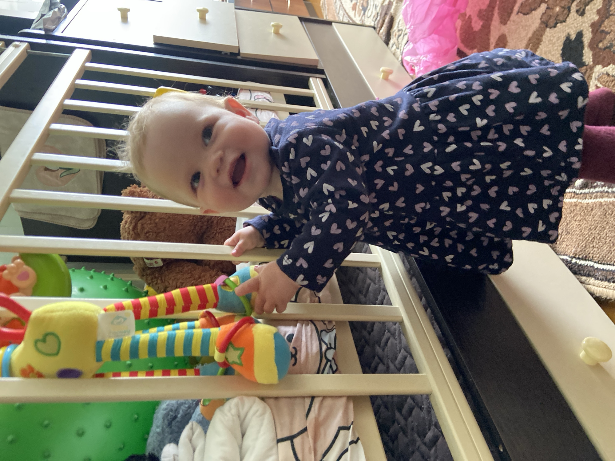 A baby girl is lying in a crib filled with toys. The baby is wearing a dark blue dress with white hearts and red tights. She is smiling and looking at the camera, holding a colorful plush toy. The crib is white and made of wood. It contains several toys, including a green spiky ball, a brown teddy bear, and other plush toys. The crib is set up in a room with a wooden floor and a rug. The background includes a light-colored cabinet with drawers and a pink tutu. The location cannot be verified as Фоки, Russia based on the image.