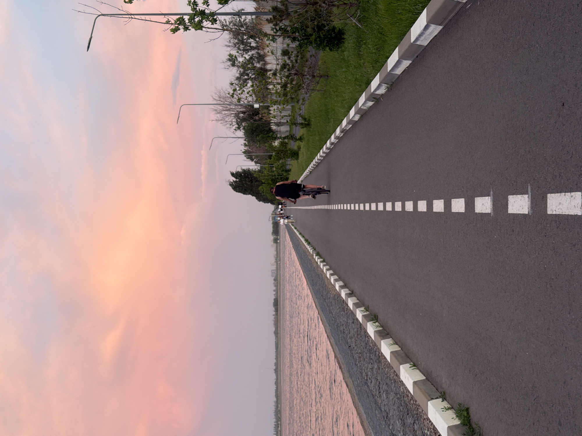 The image depicts a paved path, likely a bike path or promenade, bisected by a dashed white line. A person is riding a bicycle along the path, moving away from the viewer. A row of street lights lines the path on the left, with green vegetation and trees behind them. On the opposite side of the path is a low wall or embankment. Beyond the wall is a body of water. The sky is visible in the background, displaying a soft, pastel-colored sunset. The location is in Kuleshovka, Russia.